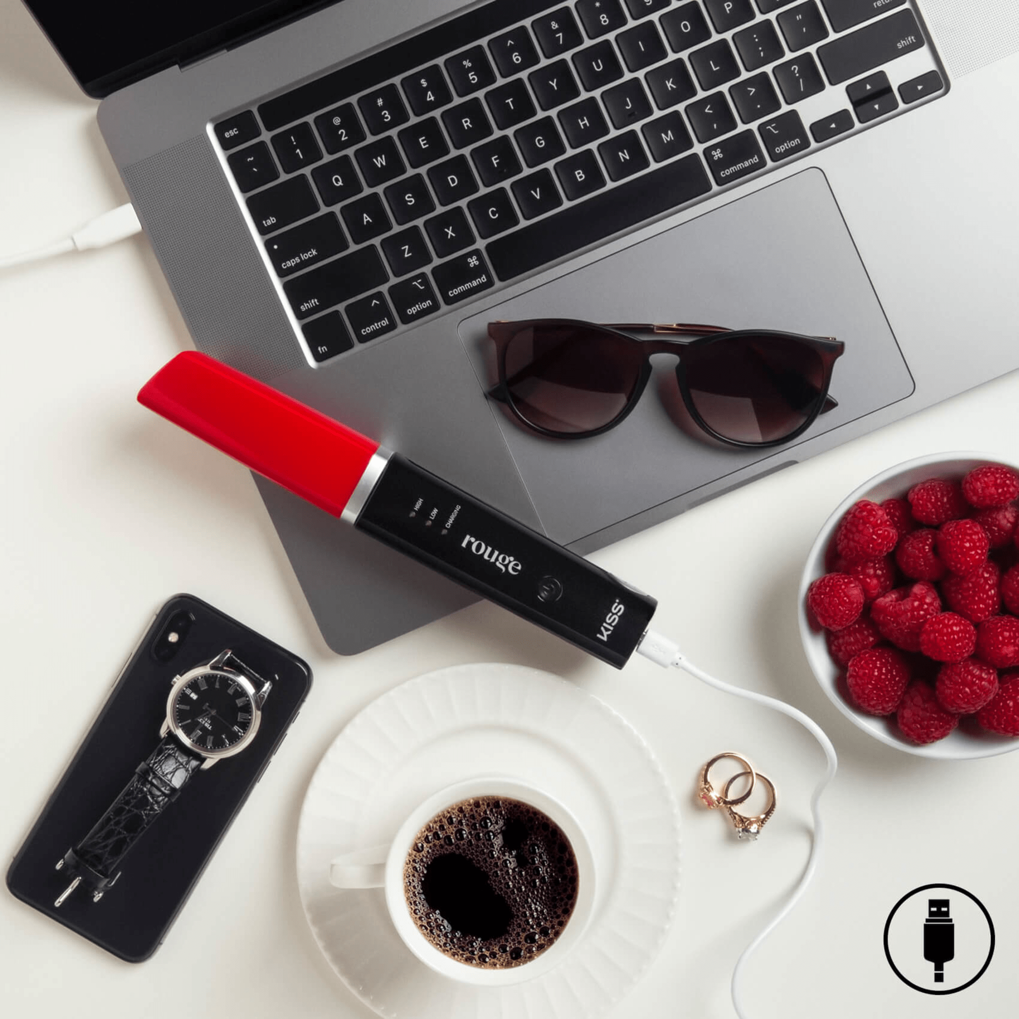 KISS portable cordless flat iron with red and black design charging via USB on a workspace with laptop, sunglasses, phone, watch, coffee, raspberries, and rings.