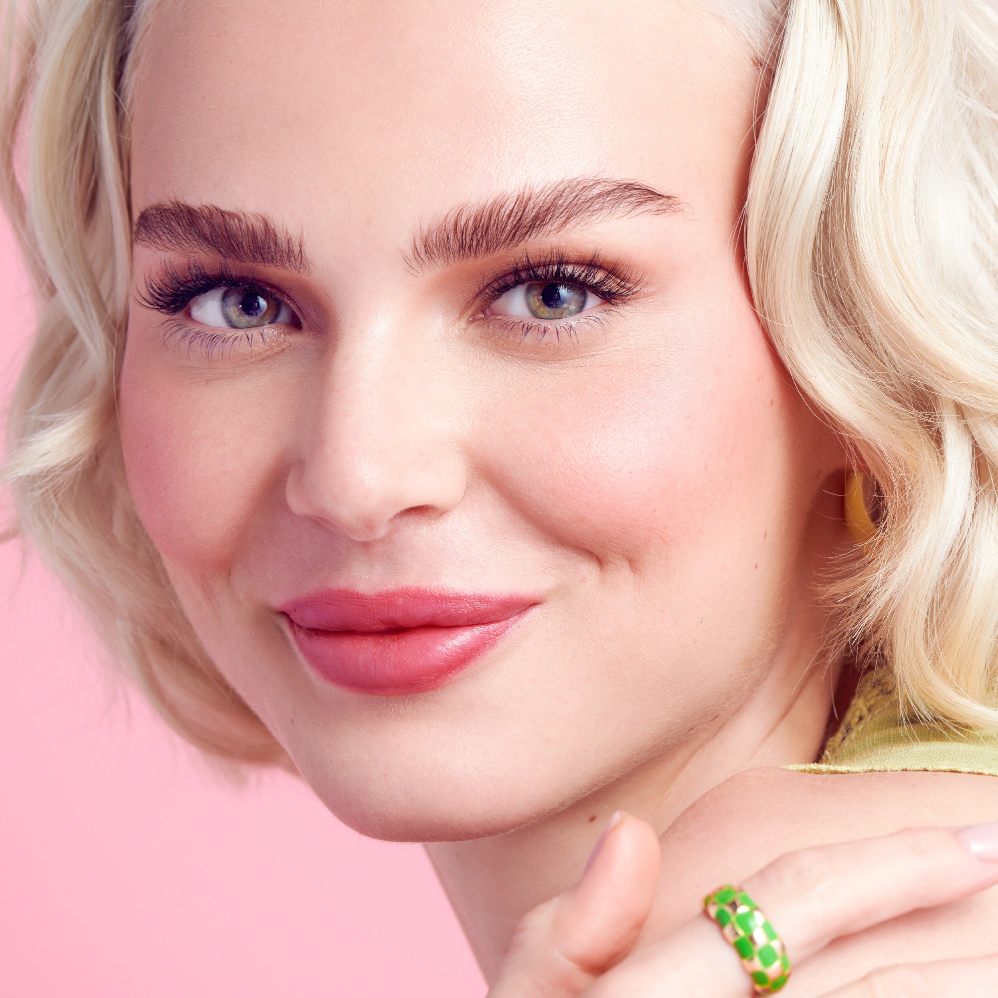 Close-up of a smiling woman with short blonde hair wearing natural-looking false eyelashes, soft pink lipstick, yellow hoop earrings, and a green checkered ring against a pink background.