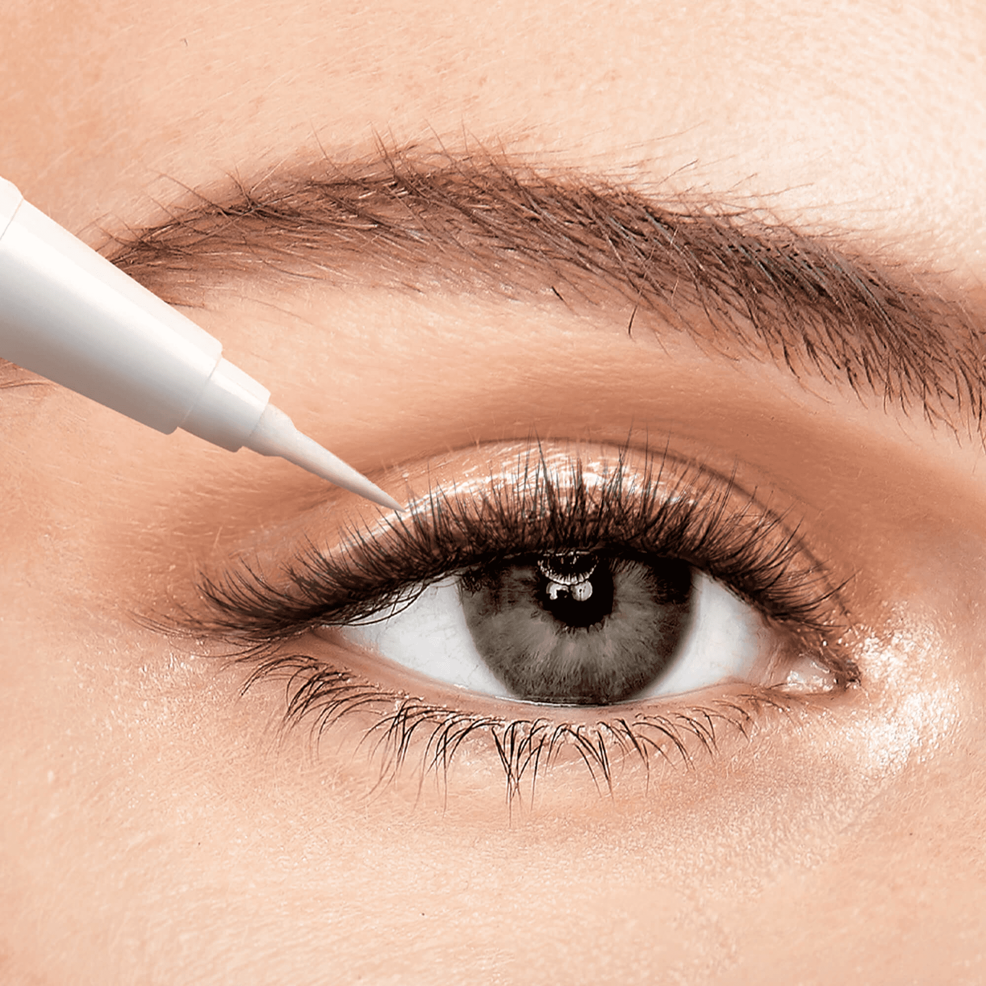 Close-up of a gray eye with long, dark eyelashes, showing a white applicator tip applying clear lash glue along the upper lash line.