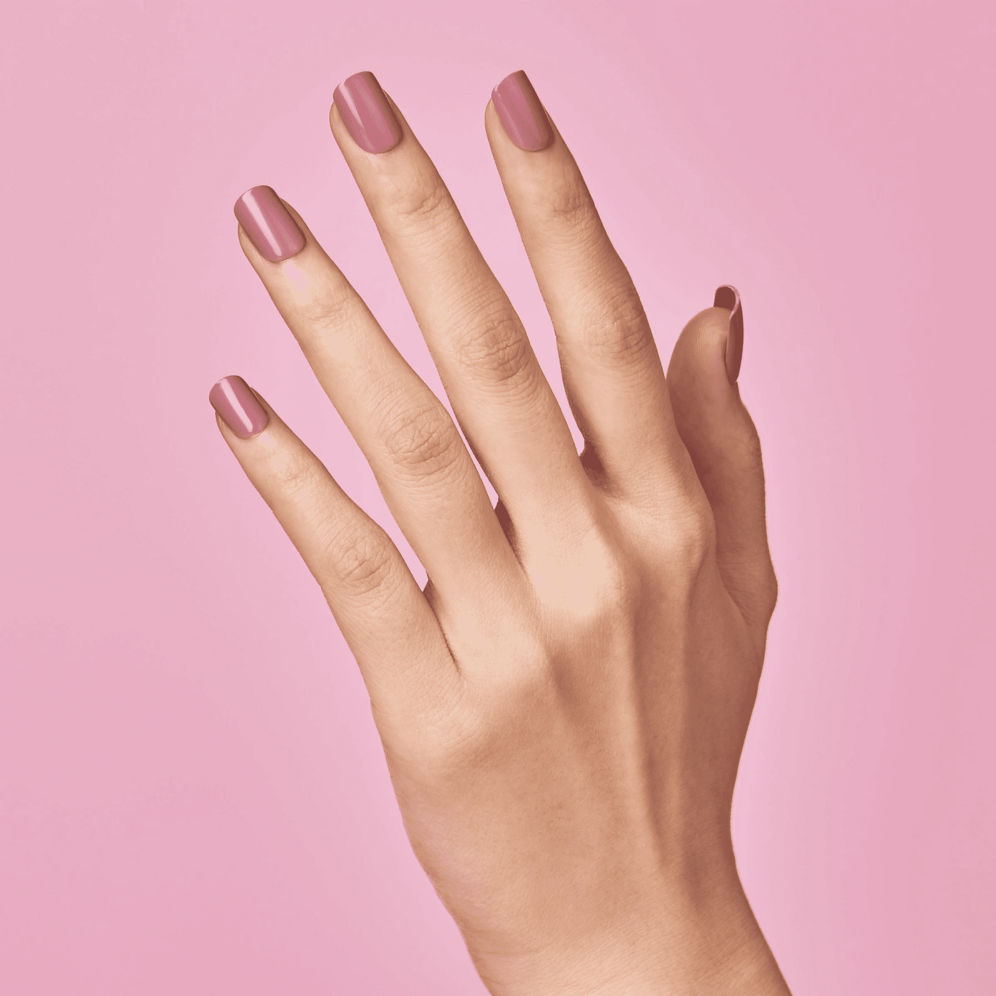 Right hand shown from the back with neatly applied, medium-length press-on nails in a muted petal pink shade against a solid pink background.