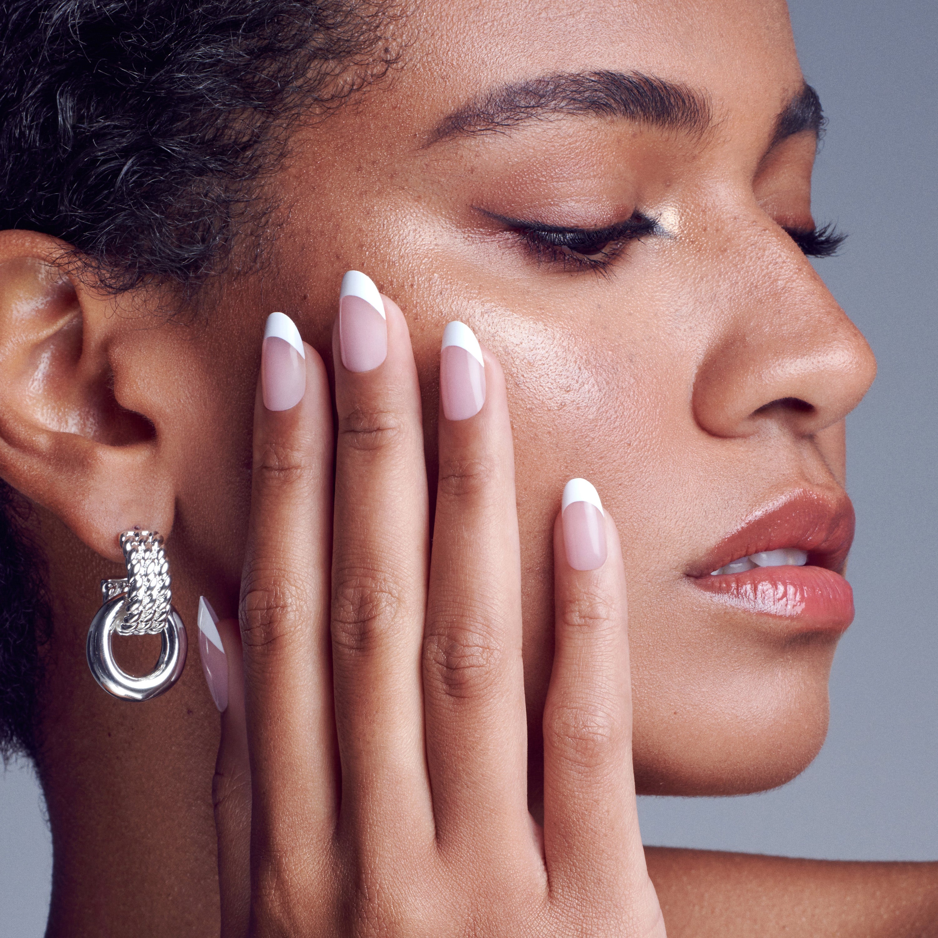 Close-up of a woman’s face with flawless makeup and glossy lips, showcasing her hand with well-groomed, natural pink nails tipped in white French manicure, wearing a textured silver hoop earring.