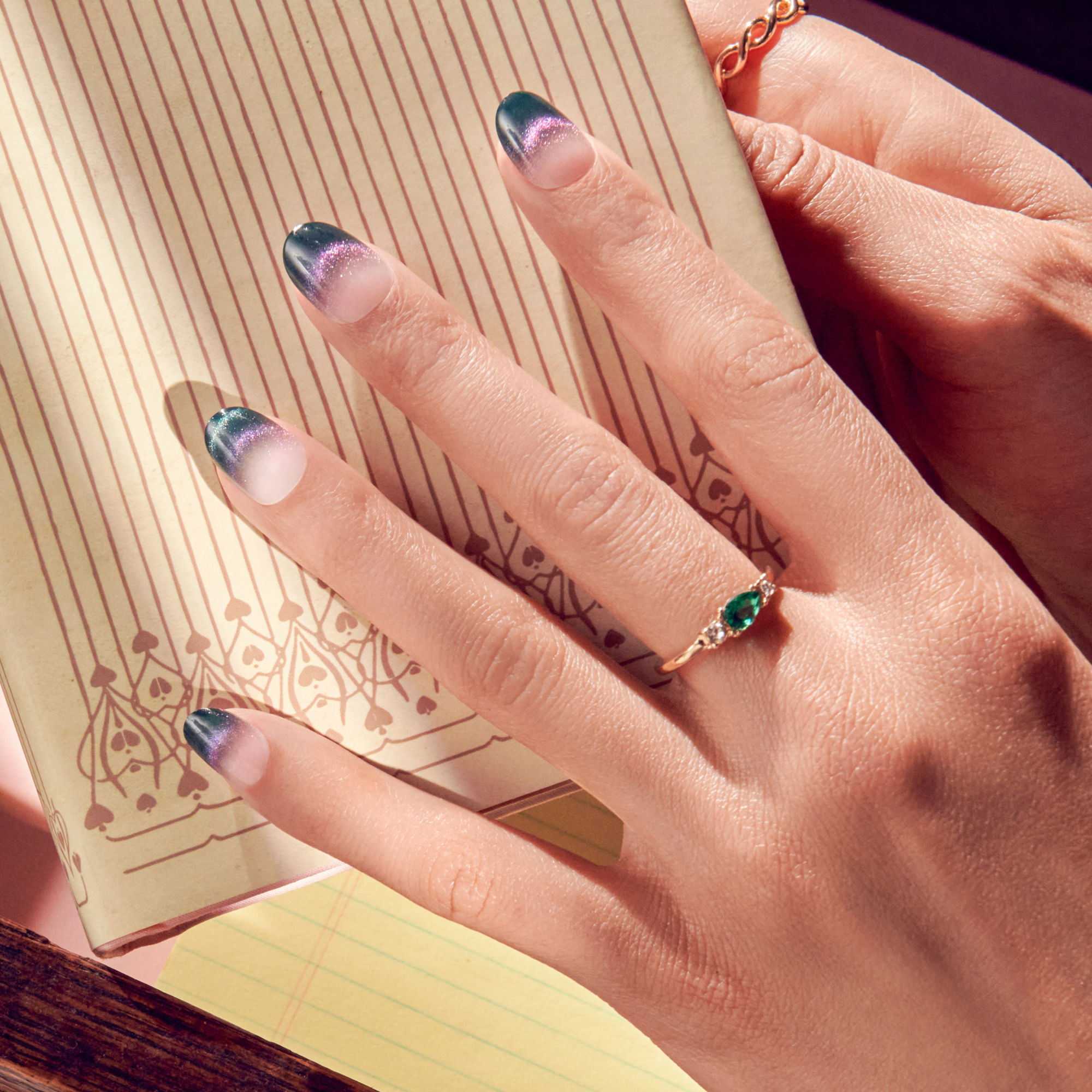 Hand with ombre navy and glitter purple French-tip nails wearing a gold ring with a green teardrop gem, resting on an illustrated beige book cover.
