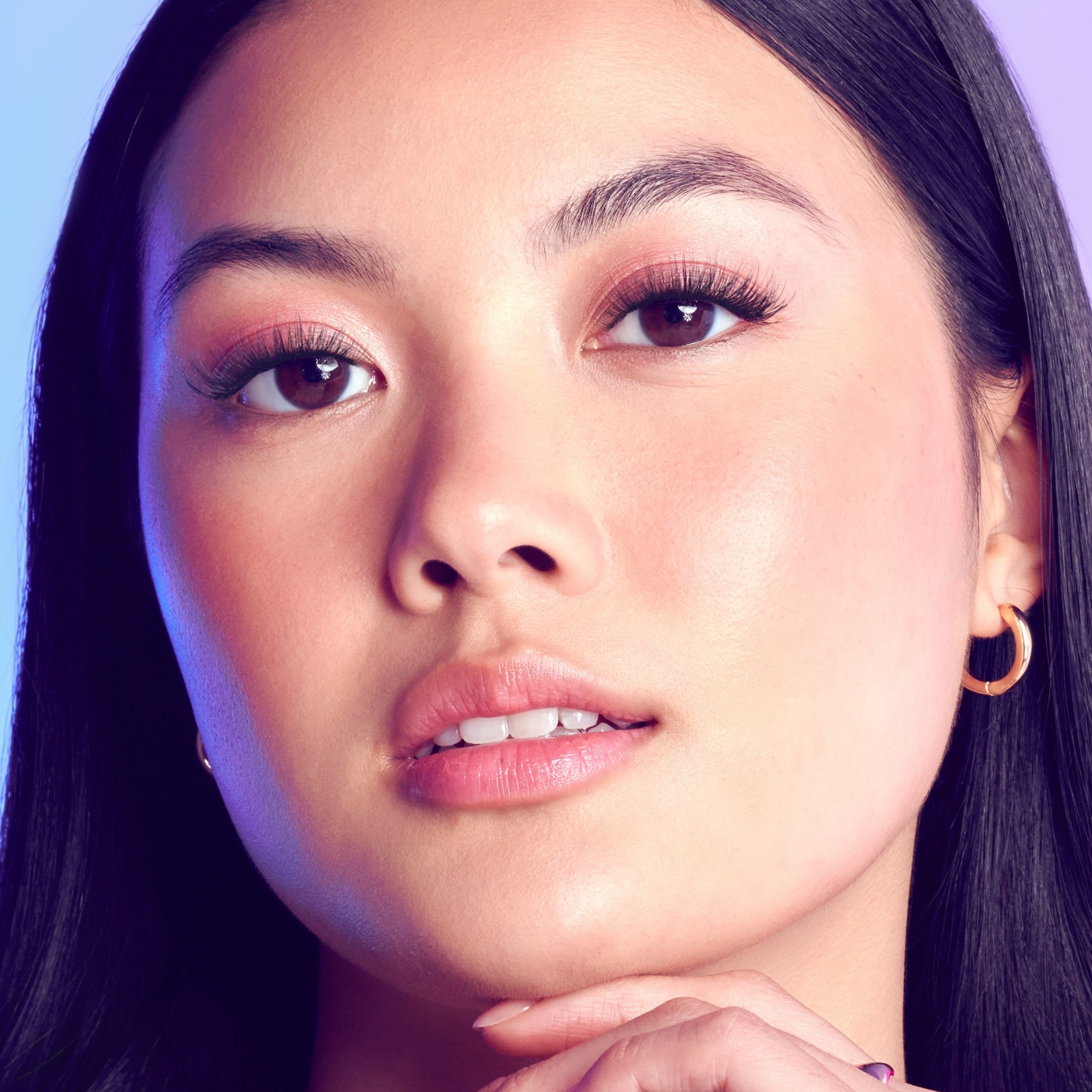 Close-up of a woman’s face showcasing long, voluminous eyelashes with natural makeup, smooth skin, and gold hoop earrings against a soft blue-purple background.