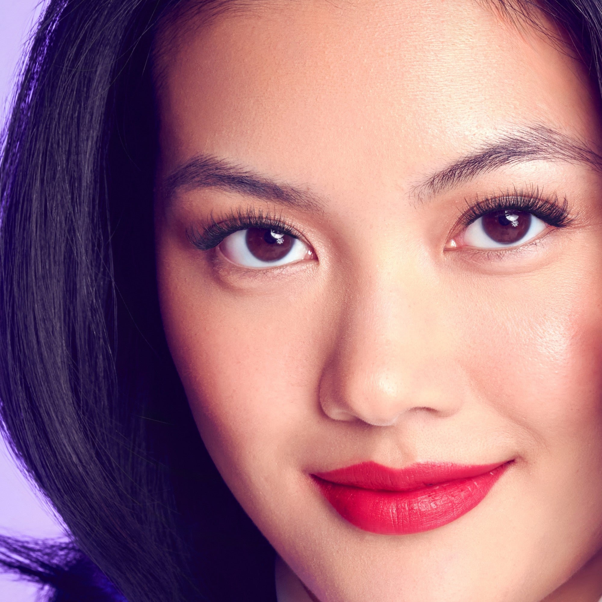 Close-up of a woman with smooth skin, glossy dark hair, bright red lipstick, and long, natural-looking false eyelashes on a purple background.