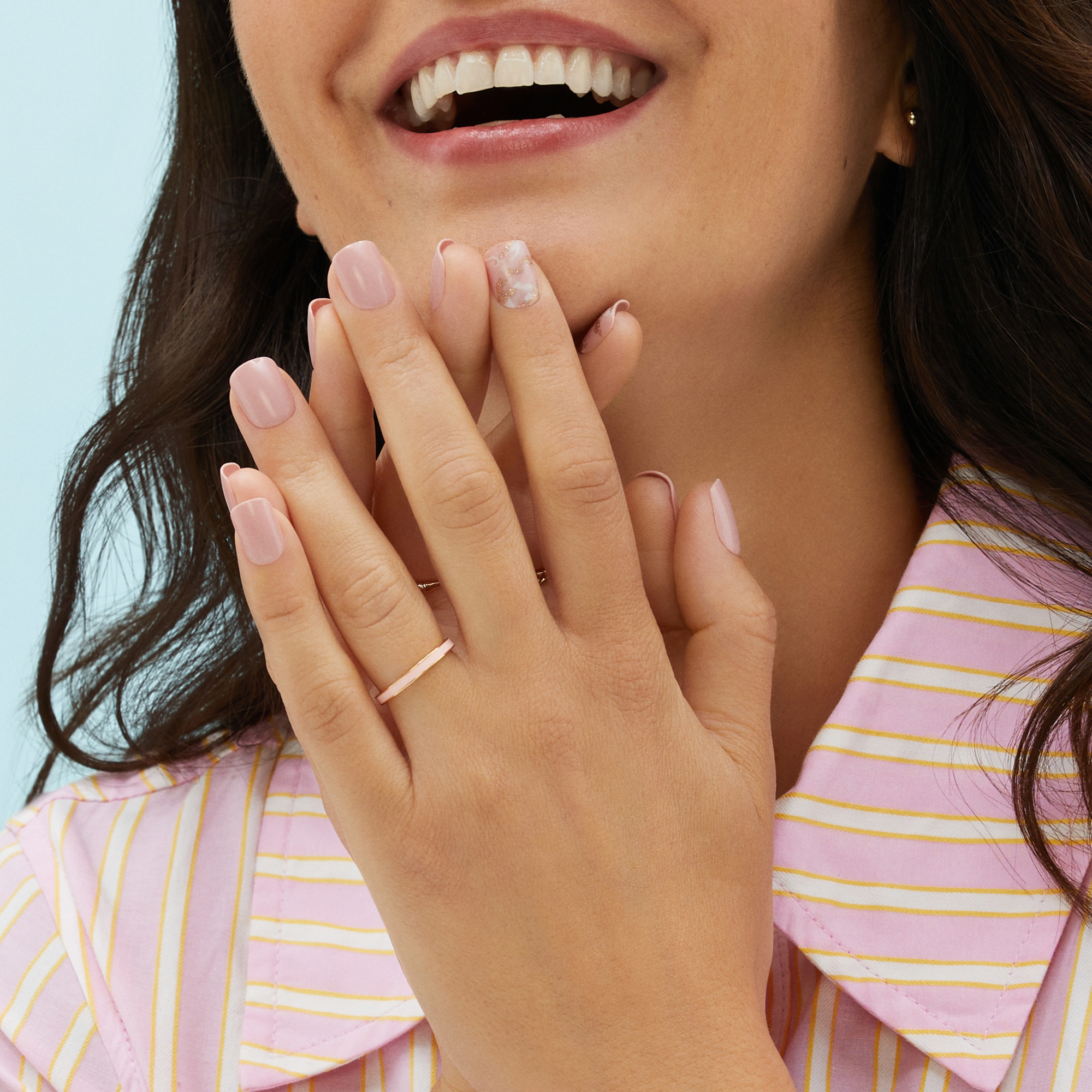 Close-up of a smiling woman wearing pastel pink press-on nails with a single accent nail featuring a pink and gold marble design, hands near her face.