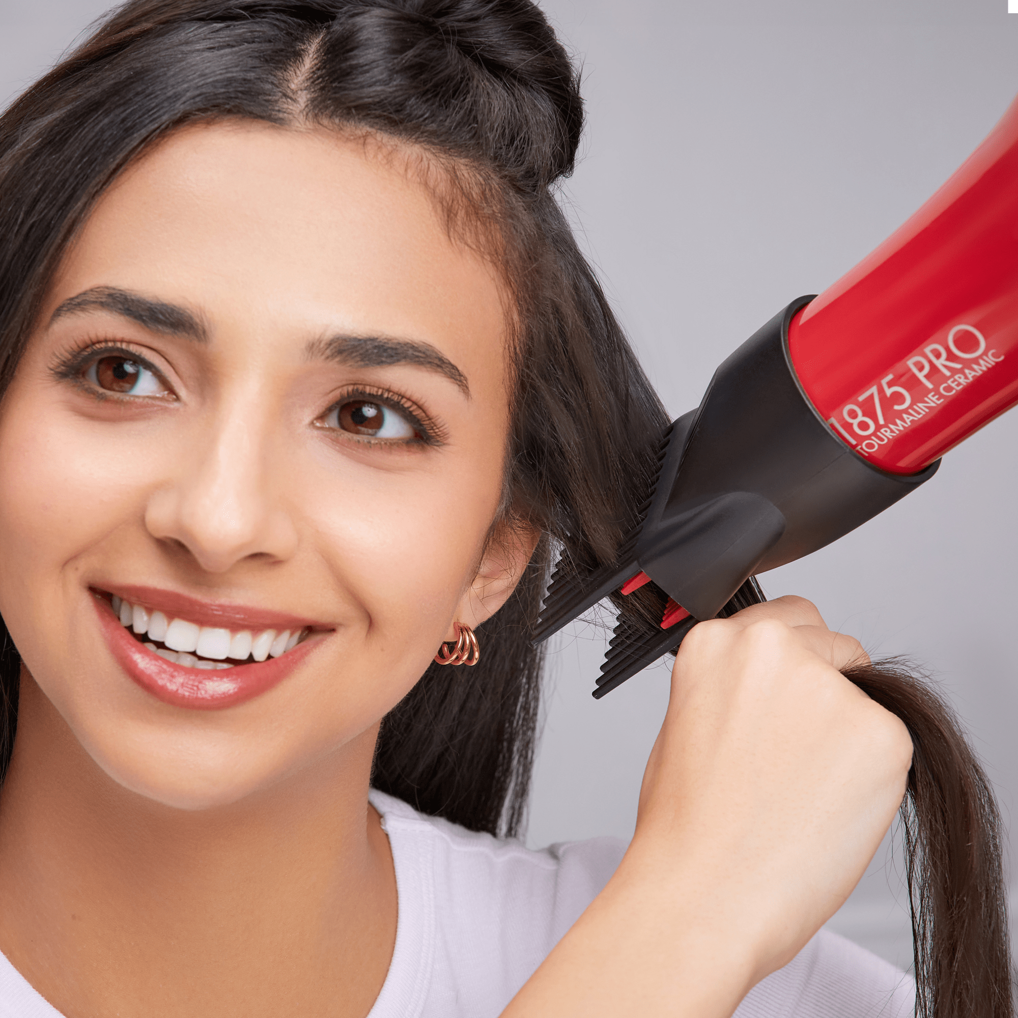 Woman using a red 1875 Pro Tourmaline Ceramic hair dryer with a black concentrator nozzle, smiling while drying her long dark hair.
