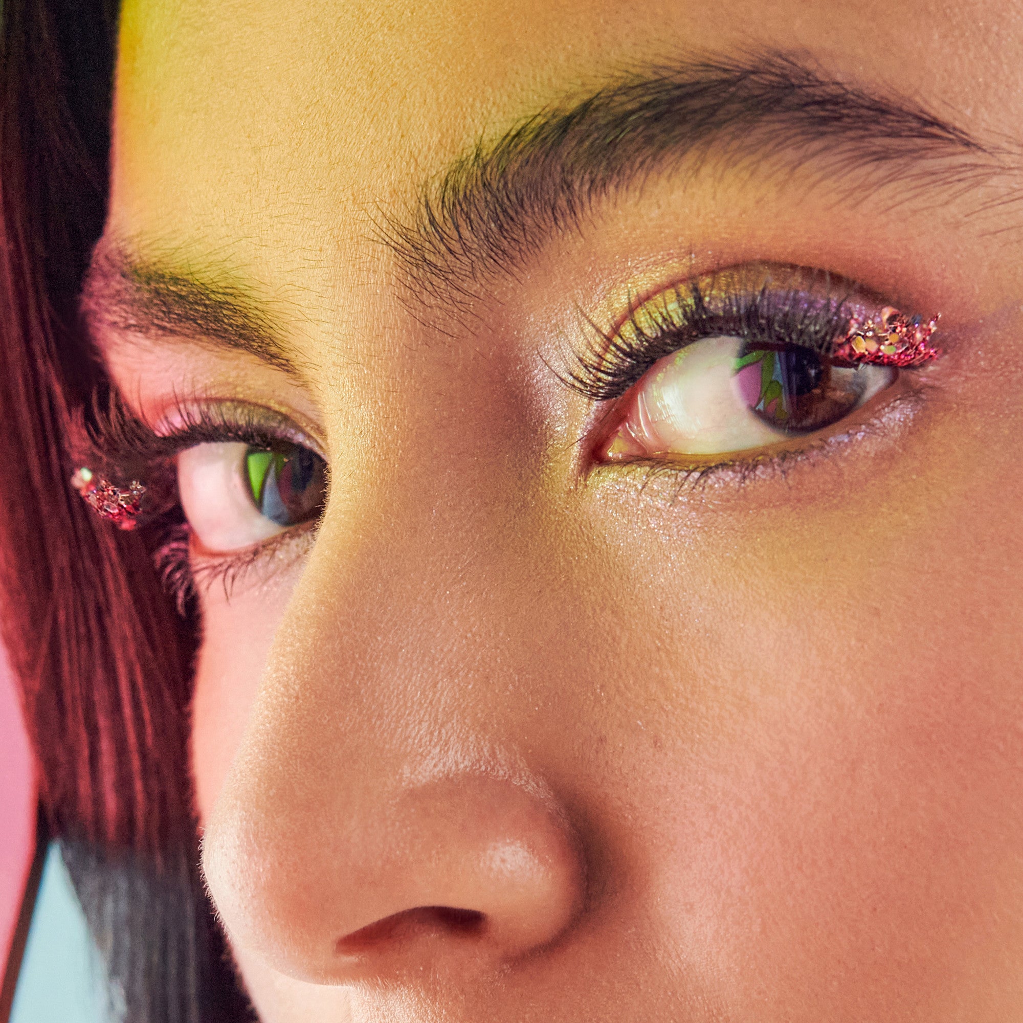 Close-up of eye makeup featuring gold eyeshadow, pink glitter liner, and burgundy hair against warm background.