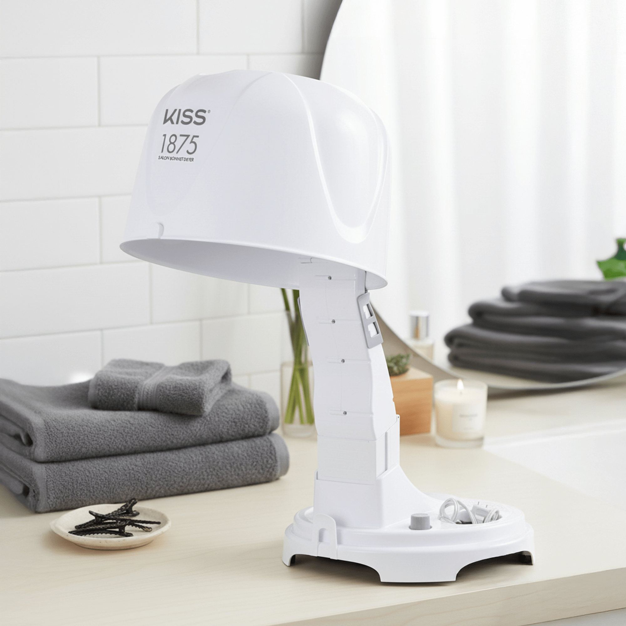 White KISS 1875W Salon Bonnet Hair Dryer on a bathroom counter with gray towels, hair clips, and a lit candle in the background.