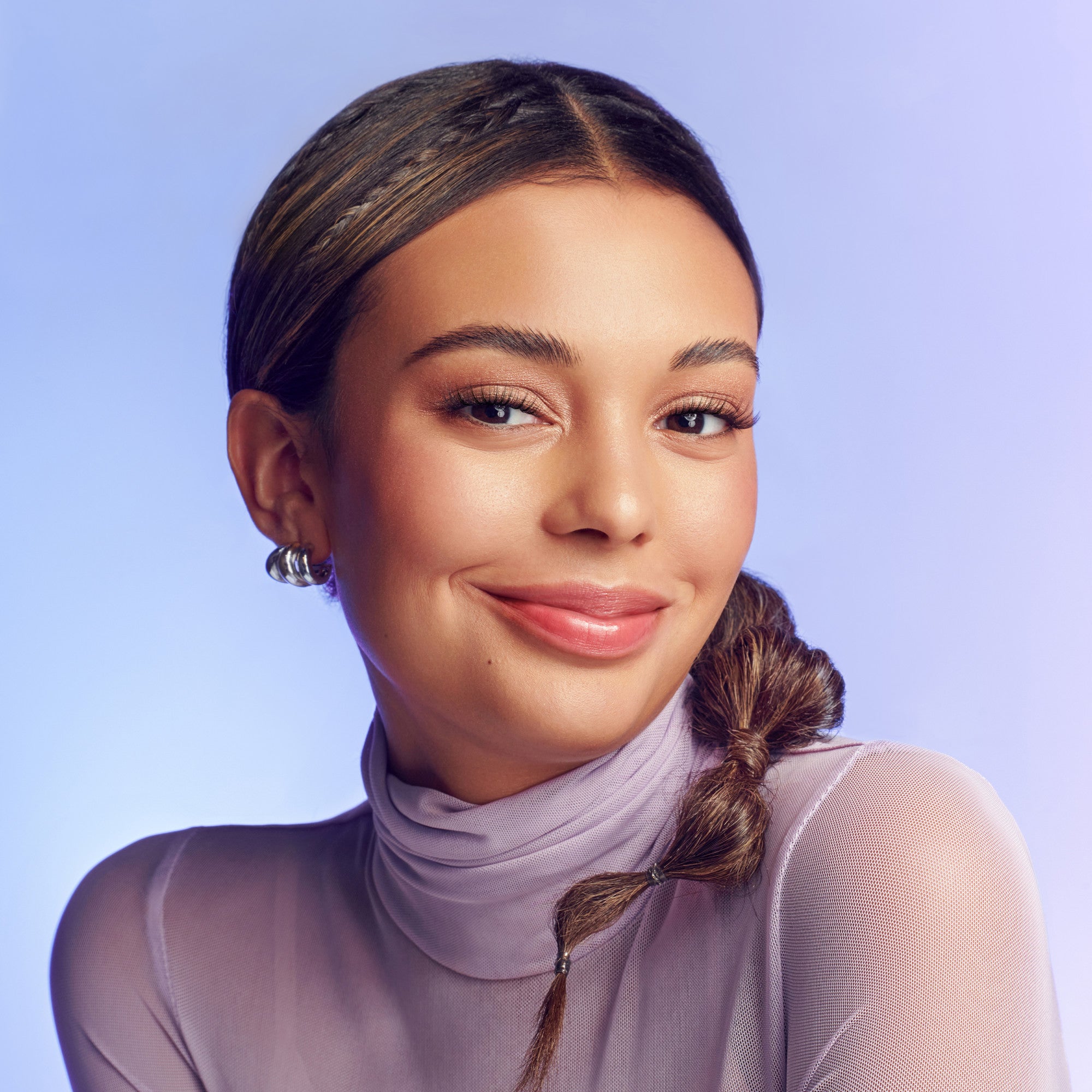 Woman with clear skin and glossy lips wearing subtle eyelash extensions and a light purple sheer turtleneck, smiling against a soft lavender background.