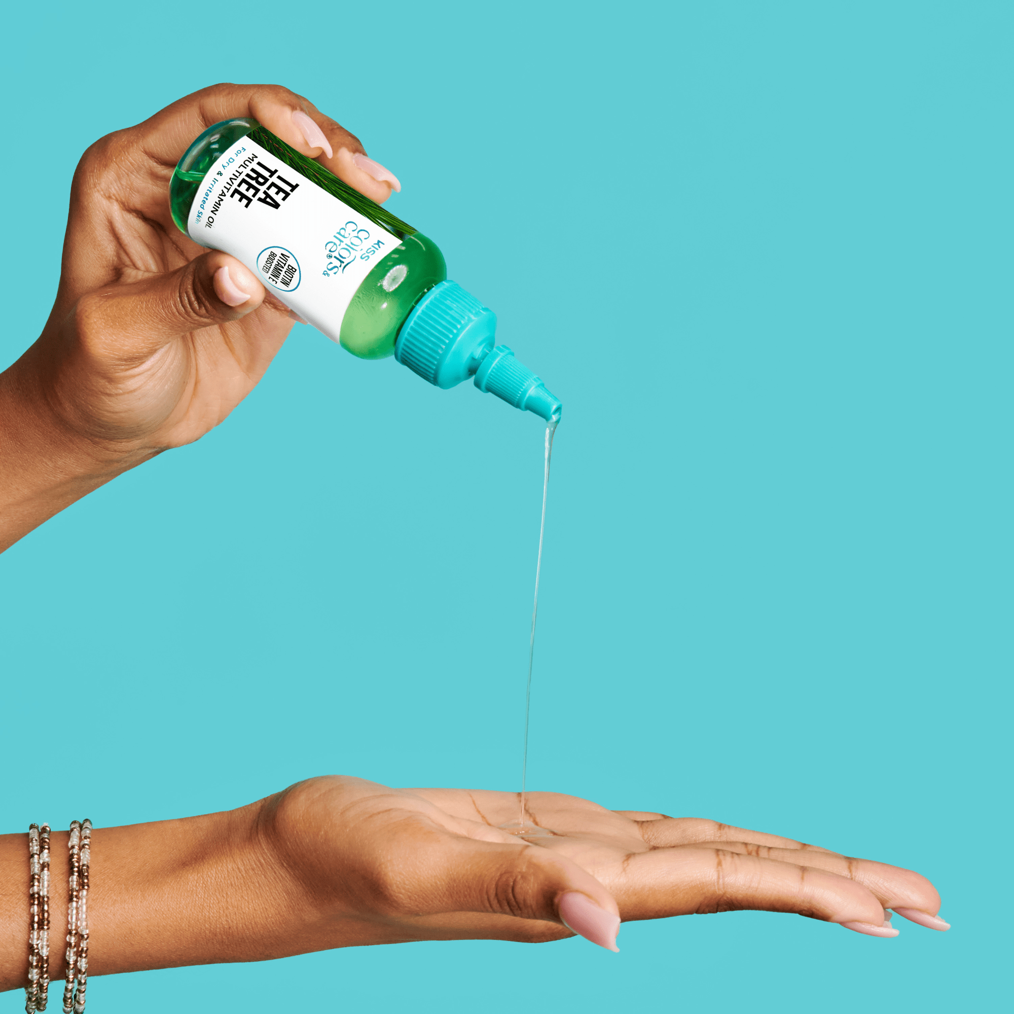 Hand dispensing clear oil from a green and white KISS Colors & Care Multivitamin Oil - Tea Tree bottle into an open palm against a blue background.