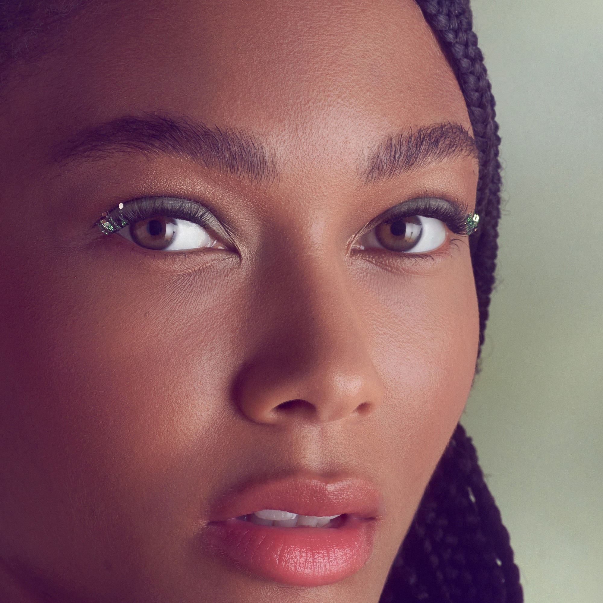 Close-up of a woman with braided hair wearing natural-looking false eyelashes with subtle green glitter accents on the outer corners.
