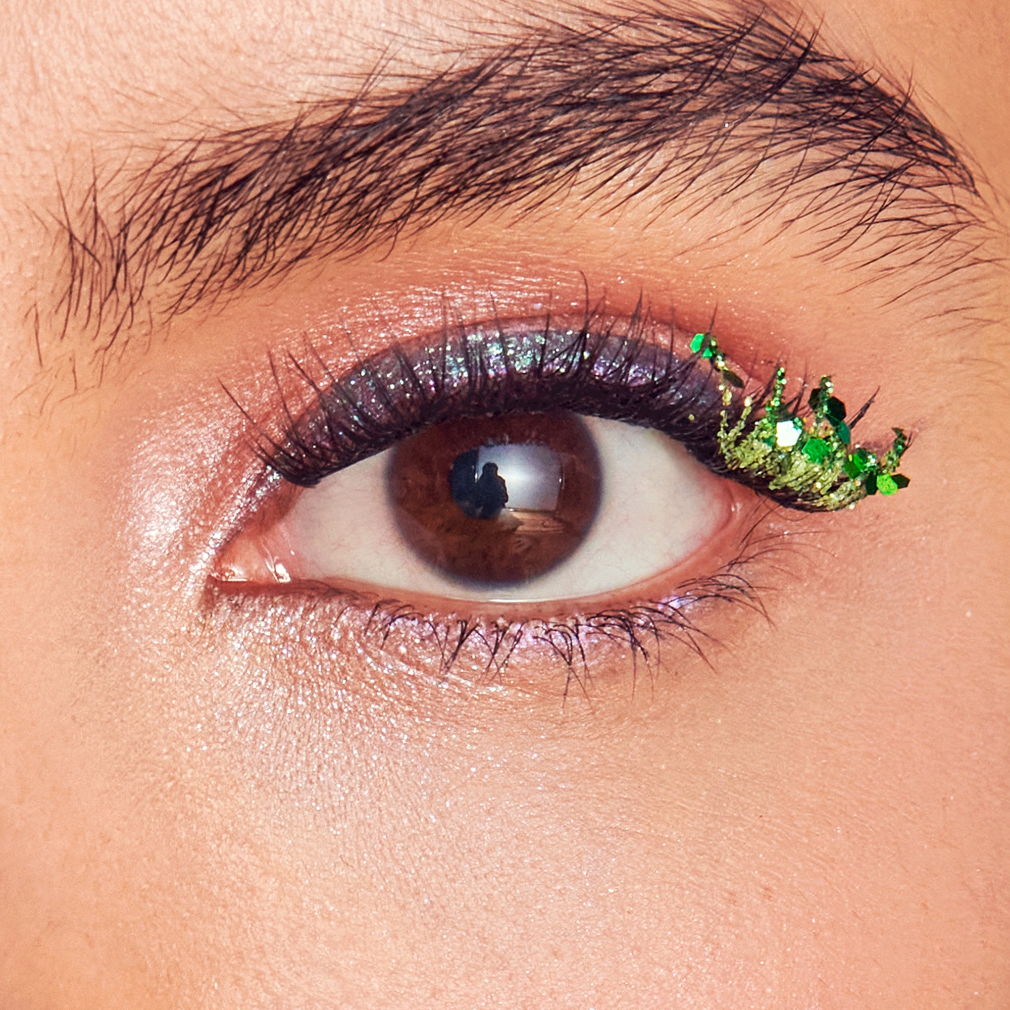 Close-up of eye with warm brown eyeshadow, black eyeliner, and green glittery decorative accent on outer corner.