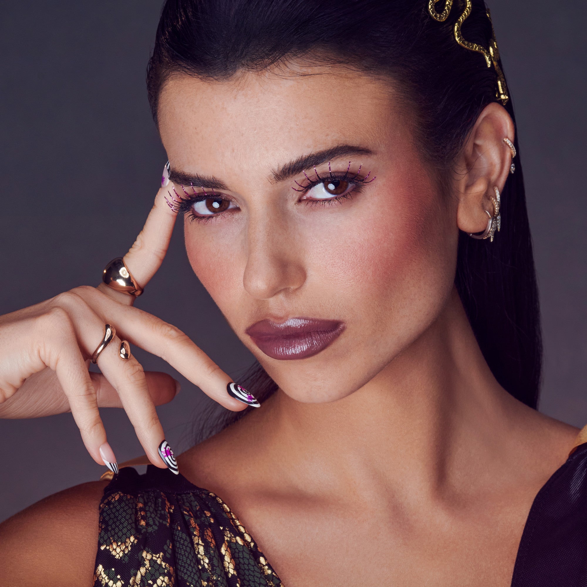 Close-up of a woman with dark hair wearing decorated false eyelashes featuring pink and black accents, complemented by bold plum lipstick, spider-themed nails, and gold jewelry.