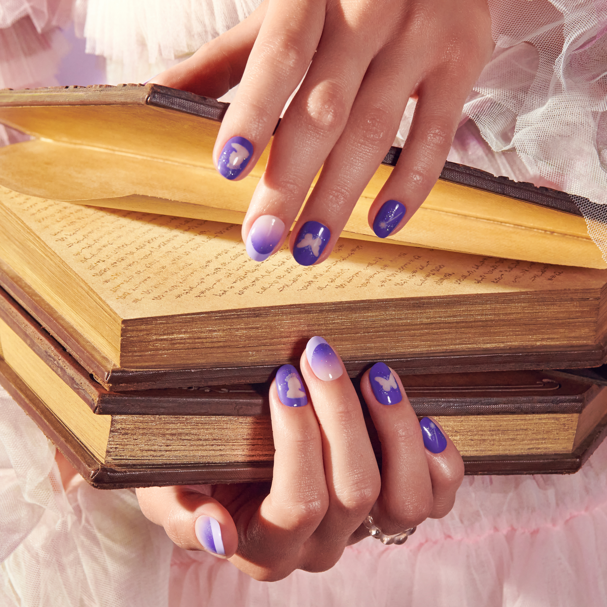 Hands with purple and white gradient nails featuring butterfly and silhouette designs, holding two stacked vintage books with yellowed pages.