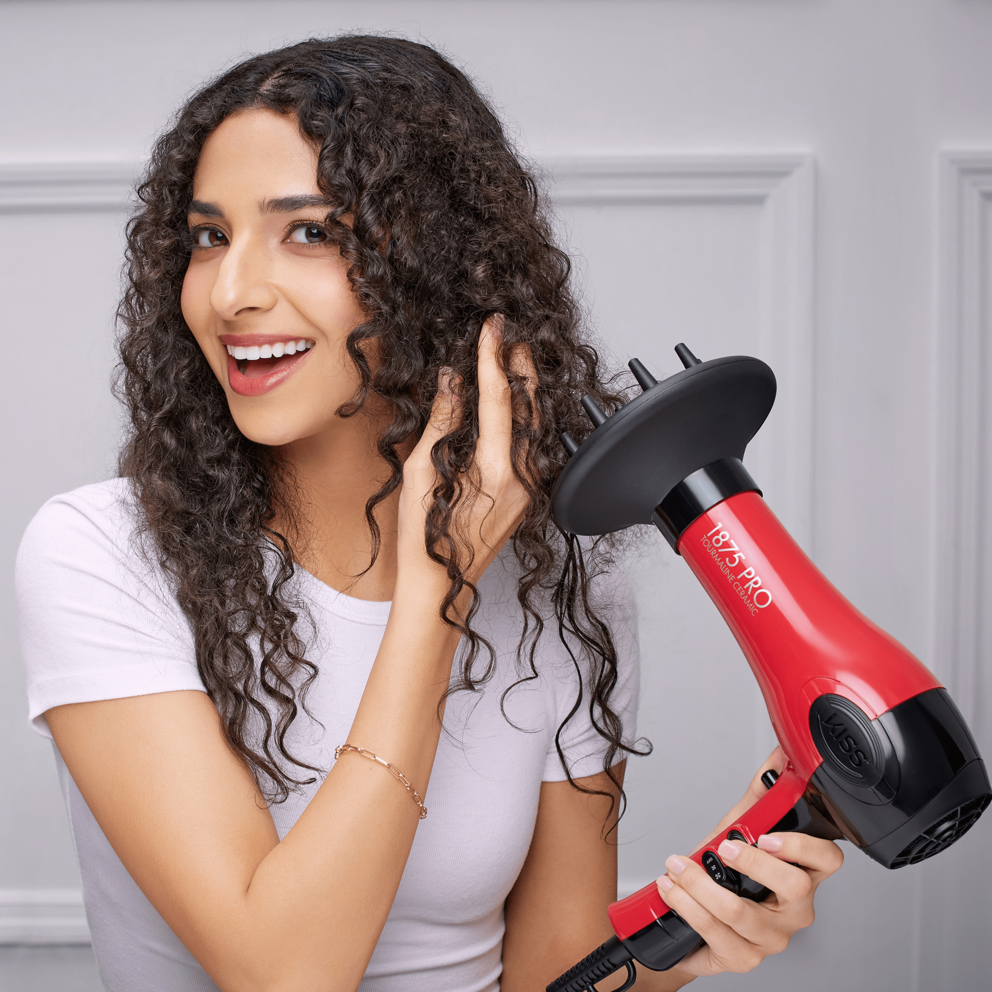 Woman with long curly hair smiling as she uses a red 1875 Pro Tourmaline Ceramic hair dryer with a black diffuser attachment in a bright indoor setting.