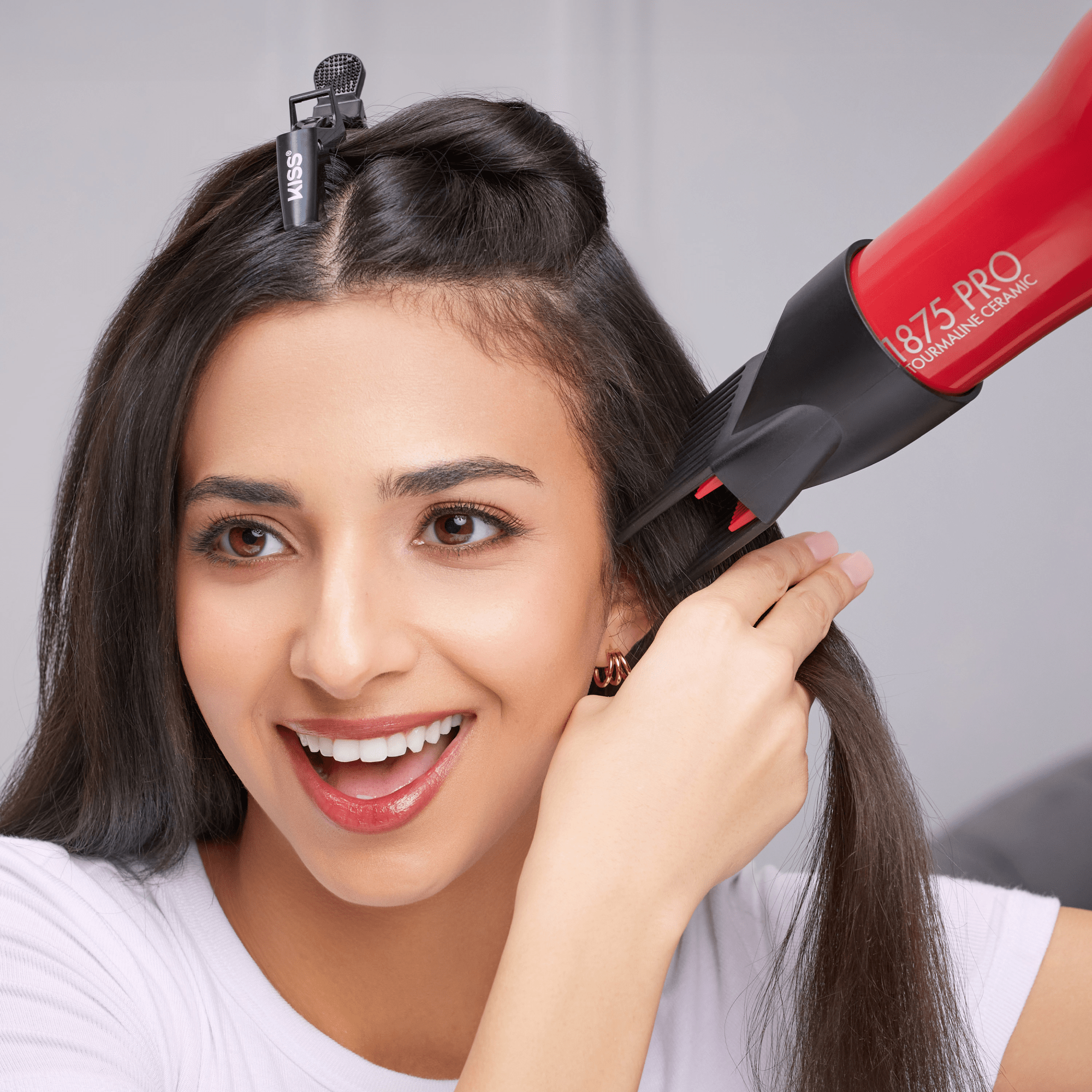 Smiling woman using a red and black 1875 Pro Tourmaline Ceramic Dryer on her straight dark hair, secured with a black KISS hair clip.