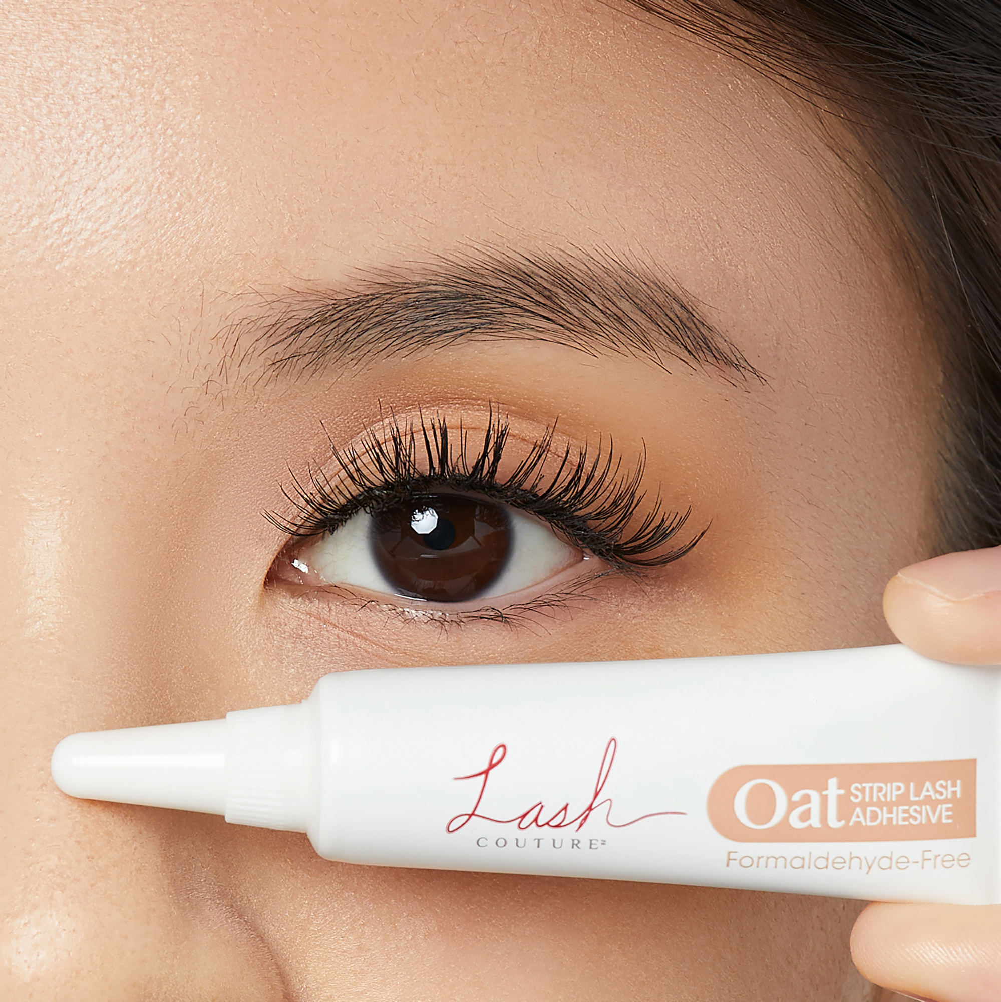 Close-up of a brown eye with long lashes holding a white tube of Lash Couture Oat Strip Lash Adhesive, labeled \