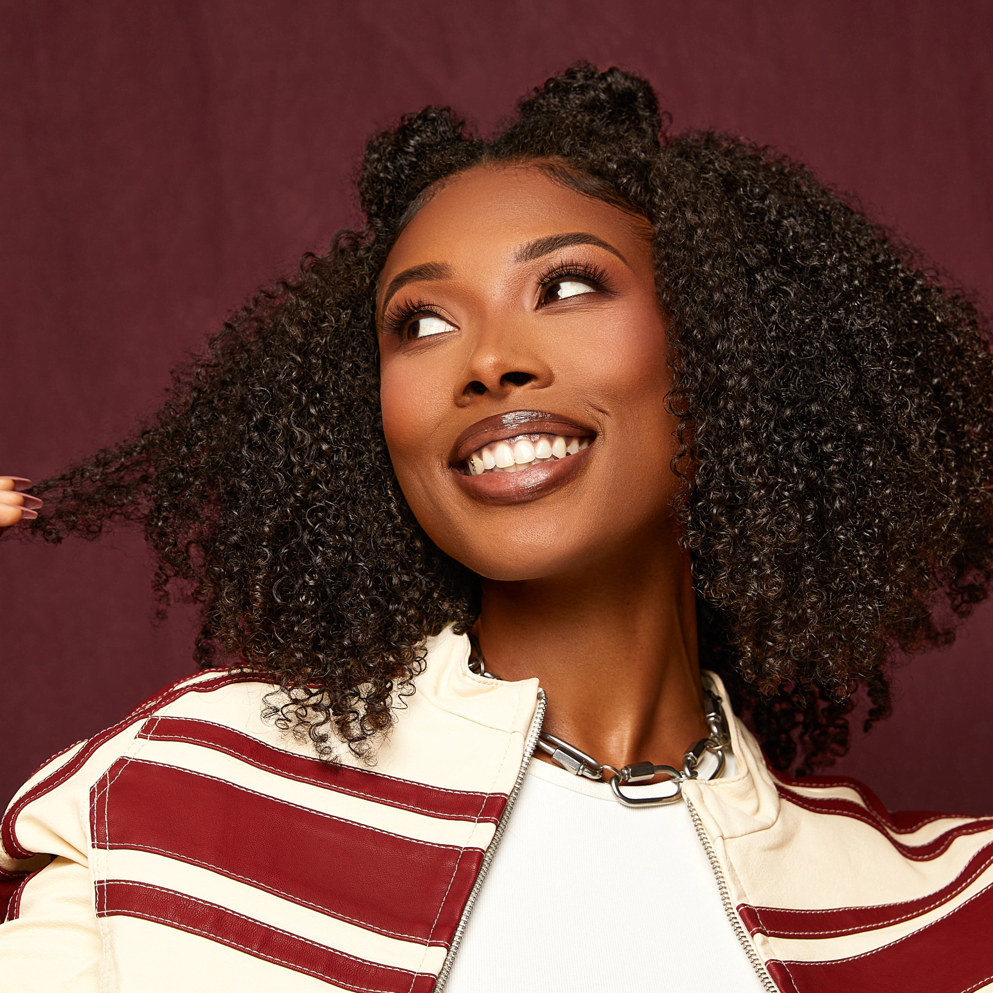 Smiling woman with shiny, healthy curly black hair wearing a white top and cream jacket with red stripes, set against a burgundy background.