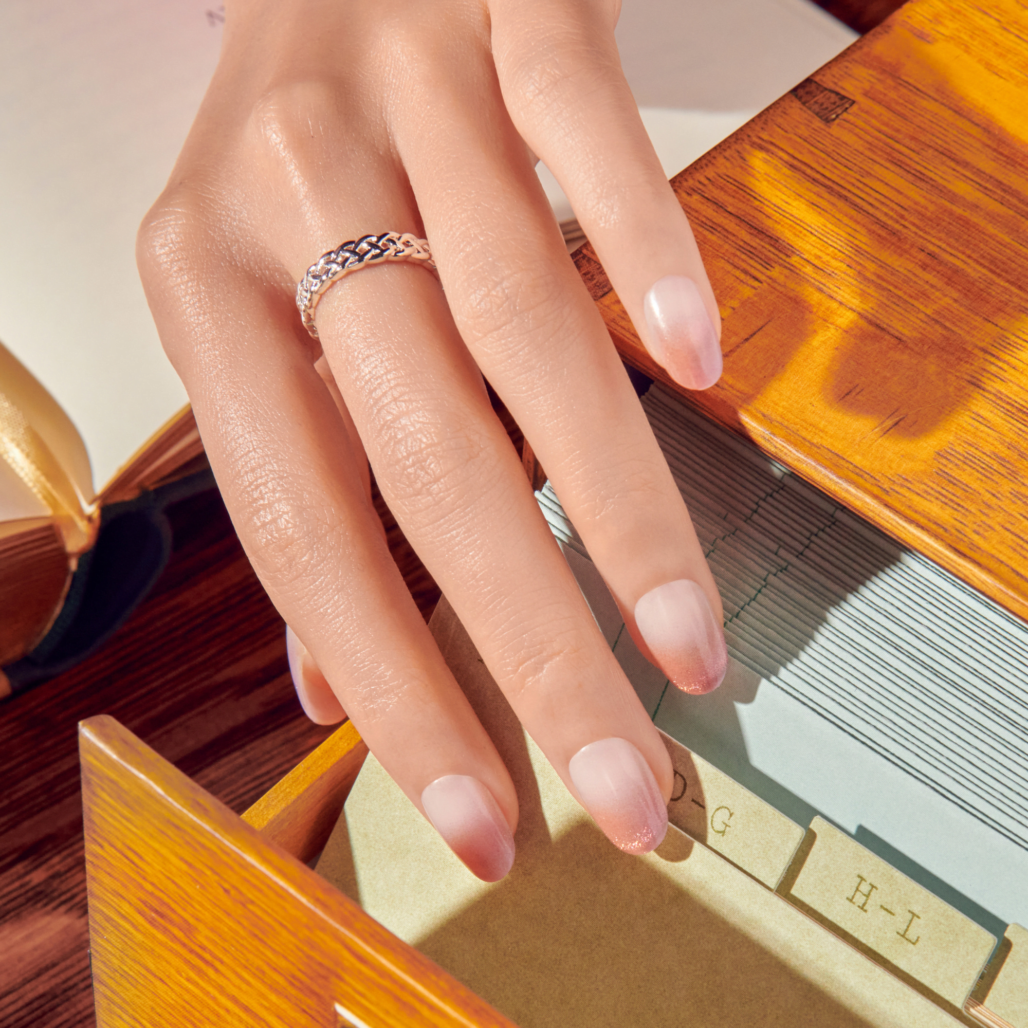 Hand with intricately braided silver ring and pink ombré nails with subtle glitter tips, resting over a wooden box and index cards with alphabetic labels.