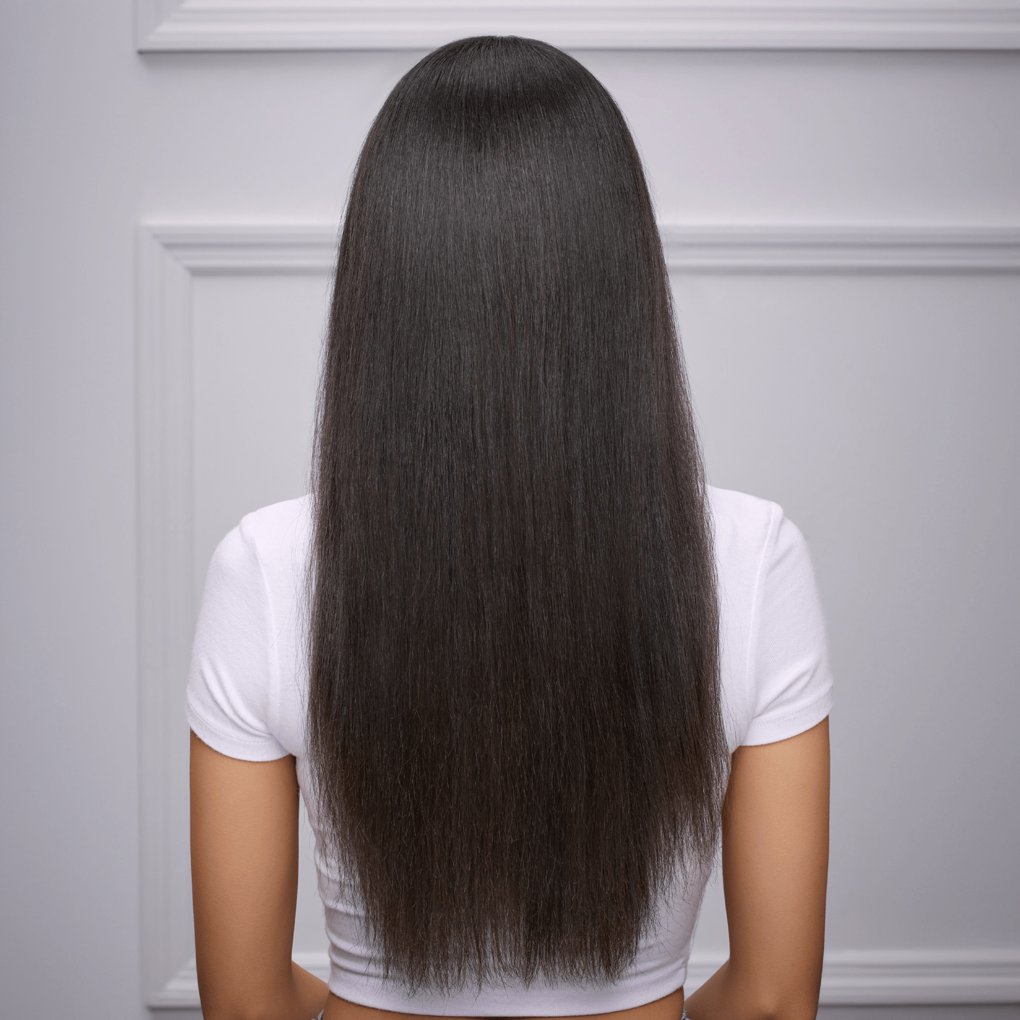 Woman with long, smooth, straight dark hair wearing a white short-sleeve top, shown from the back against a light gray wall with decorative molding.