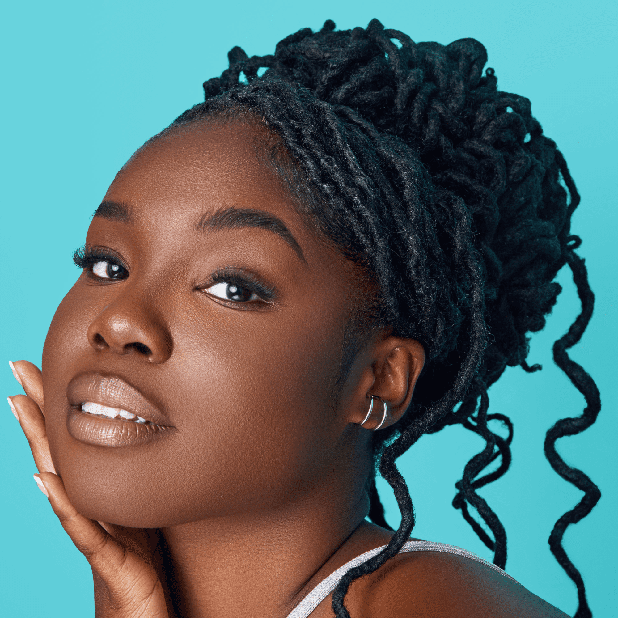 Close-up portrait of a woman with styled black dreadlocks against a bright blue background, gently touching her face with her hand.