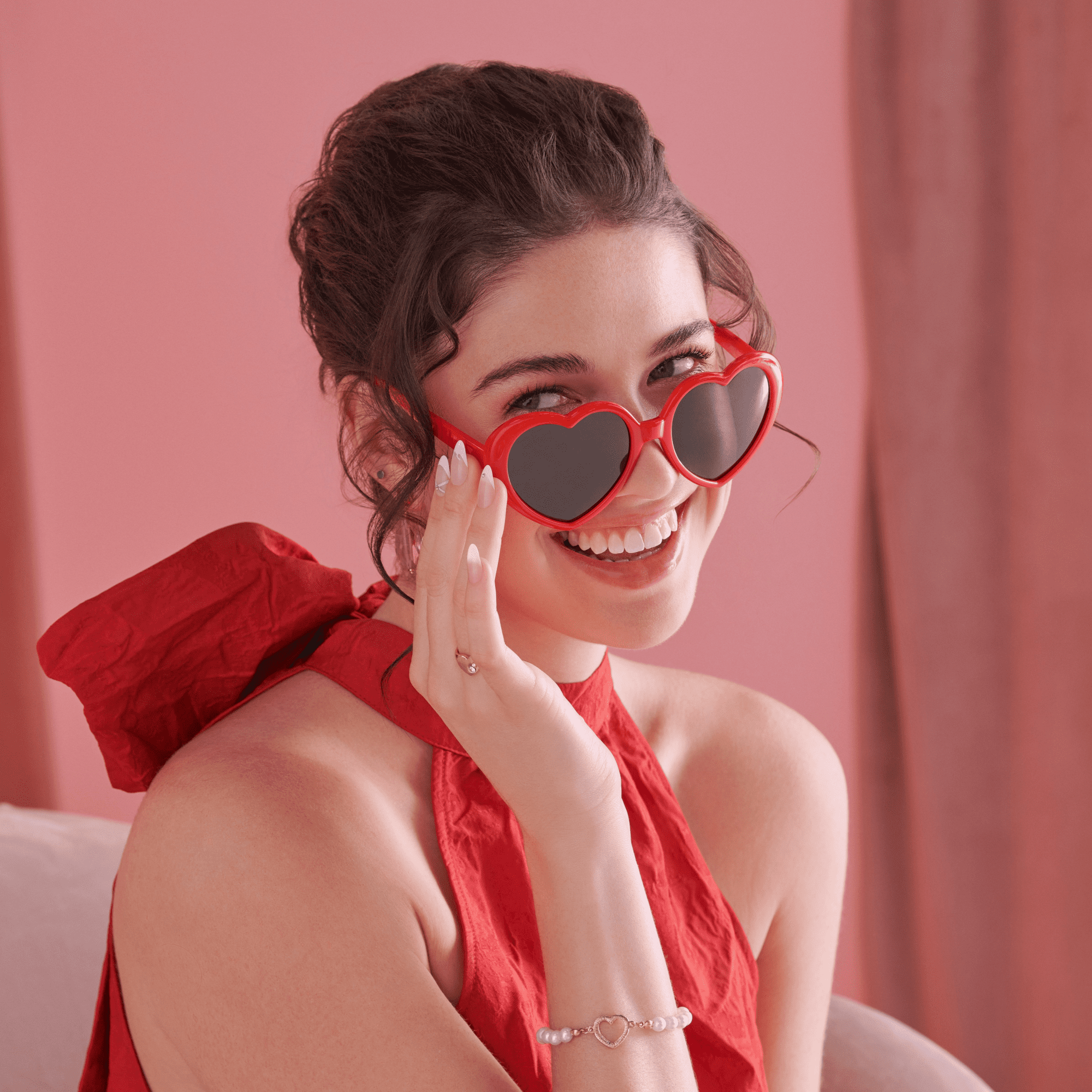 Smiling woman in a red halter dress and red heart-shaped sunglasses shows off white press-on nails with heart designs on a pink background.