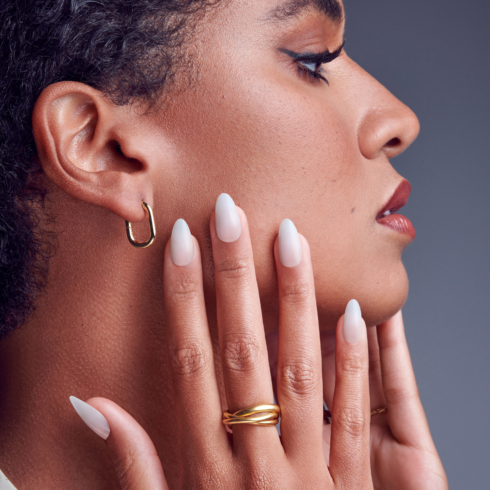 Close-up side profile of a person with short curly hair wearing gold hoop earrings and a matching gold ring, showcasing long, almond-shaped, matte white press-on gel nails.