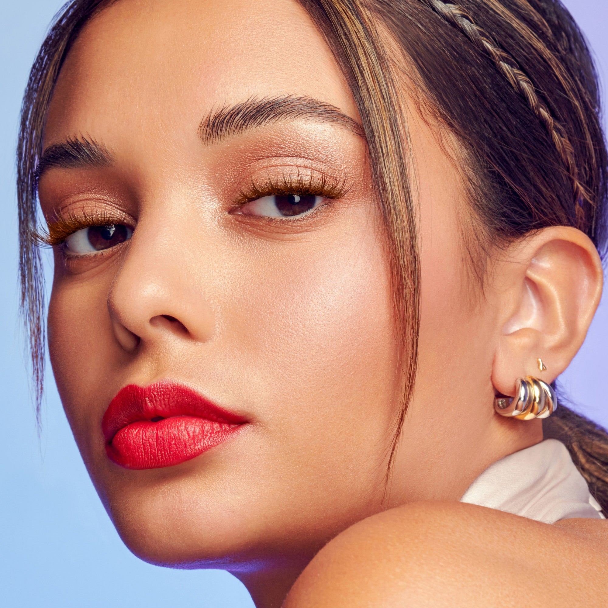 Close-up of a woman with brown wisps styled into her dark hair, highlighting natural brown eyelash extensions and warm neutral makeup against a soft blue background.