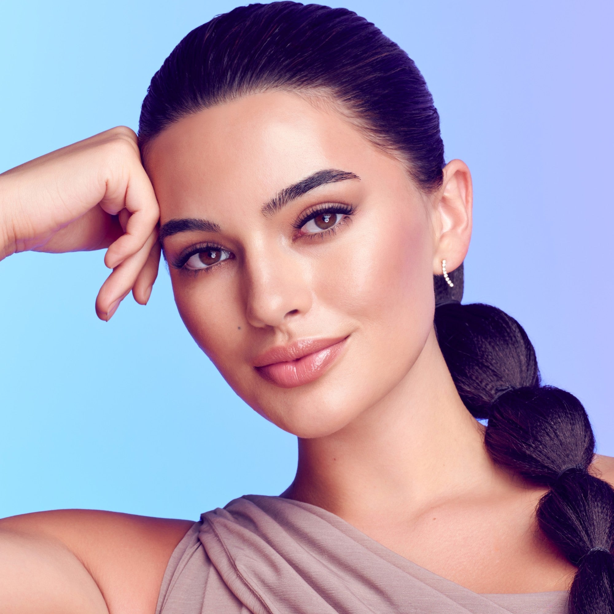 Close-up portrait of a woman with smooth skin, natural makeup emphasizing long, wispy lashes, dark braided hair, and wearing a taupe sleeveless top against a blue-purple gradient background.