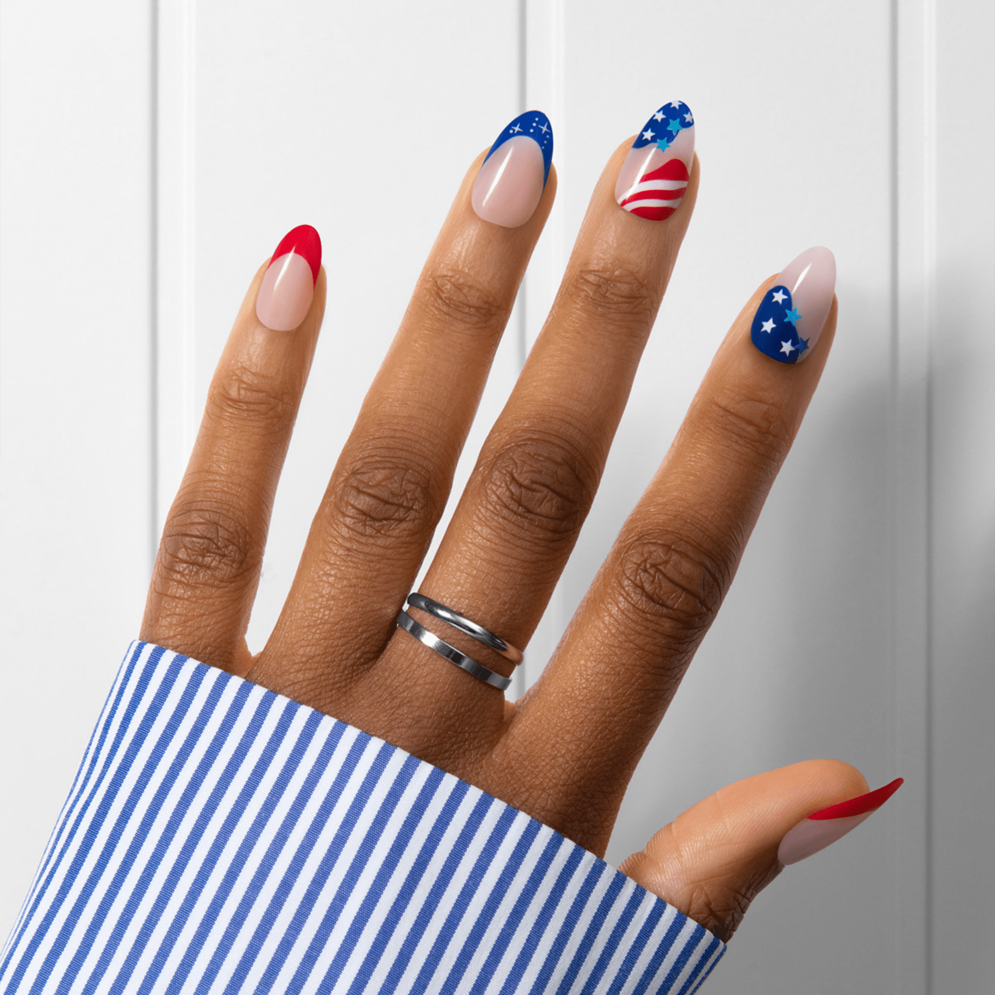 Fingernails with press-on nail design featuring red French tips, blue with white stars, and patriotic American flag-inspired patterns on a hand wearing a silver ring and blue striped shirt cuff.