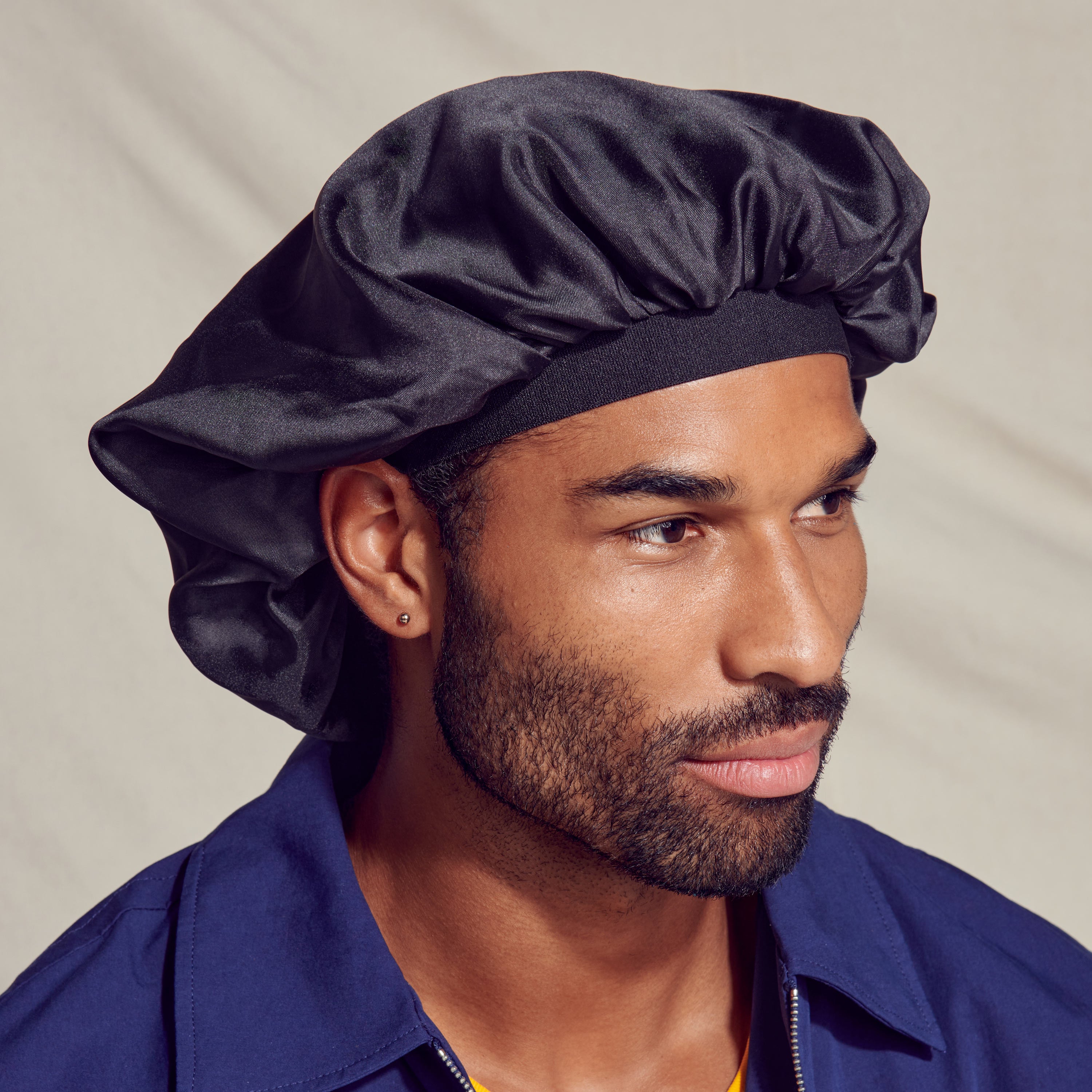 Man wearing a shiny black silky satin oversized double wear cap, shown in a close-up side view against a neutral background.