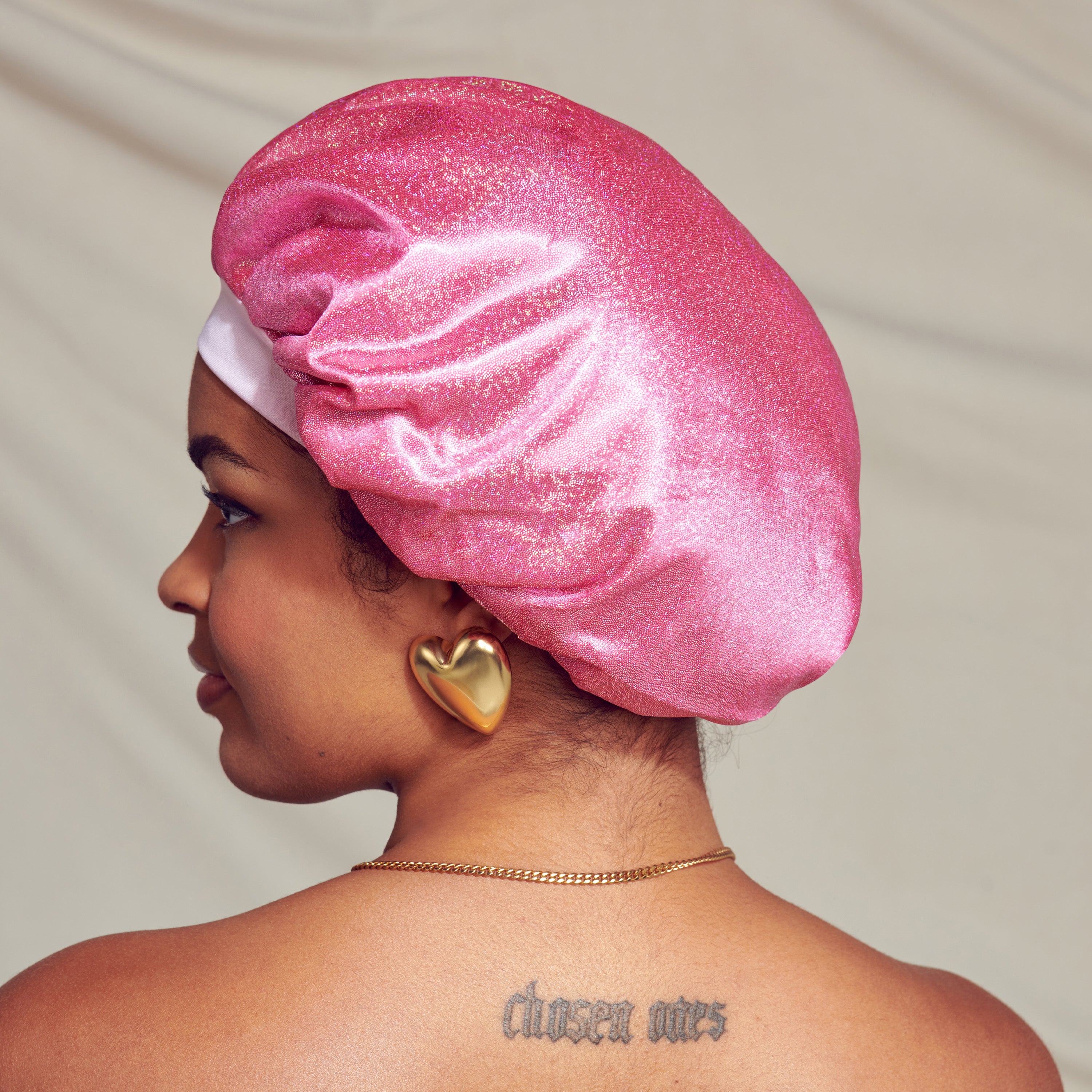 Woman shown from the side wearing a shimmery pink satin bonnet cap with glitter, styled as a slouchy hair cover, against a neutral background.
