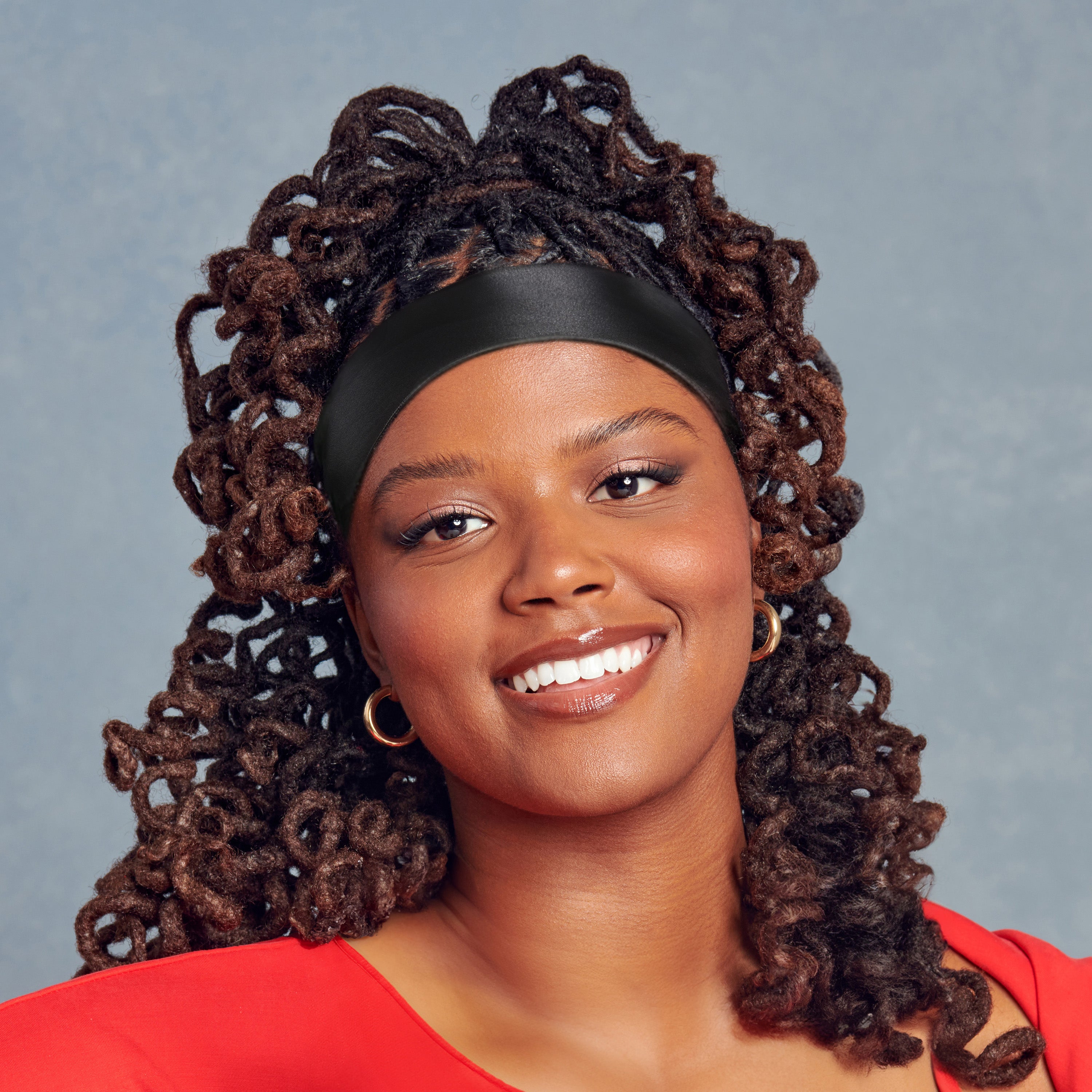 Woman with curly hair wearing a smooth black satin edge scarf styled as a wide headband, smiling against a soft gray background.