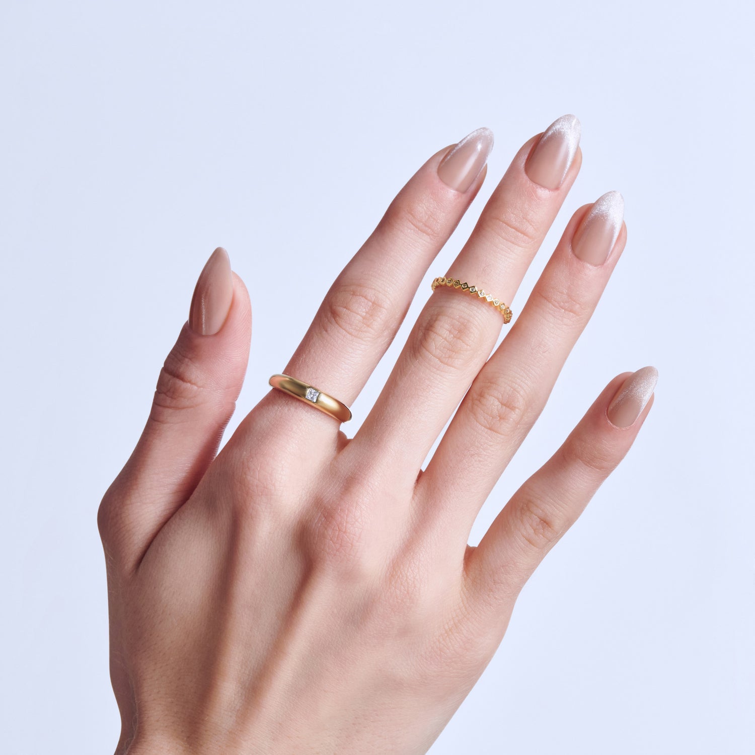 Hand displaying manicured nails with gold and diamond rings against light background.