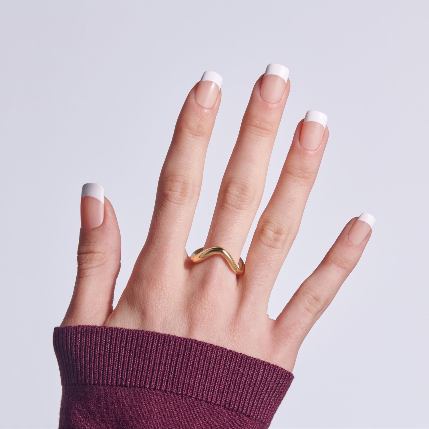 Hand with classic French manicure nails wearing gold curved band ring, burgundy sleeve visible.