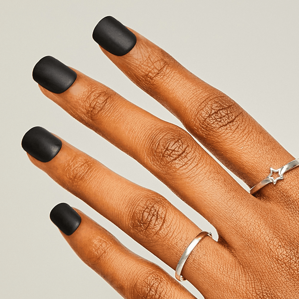 Hand with medium brown skin tone displaying matte black press-on nails in a short square shape, worn on four fingers adorned with thin silver rings.