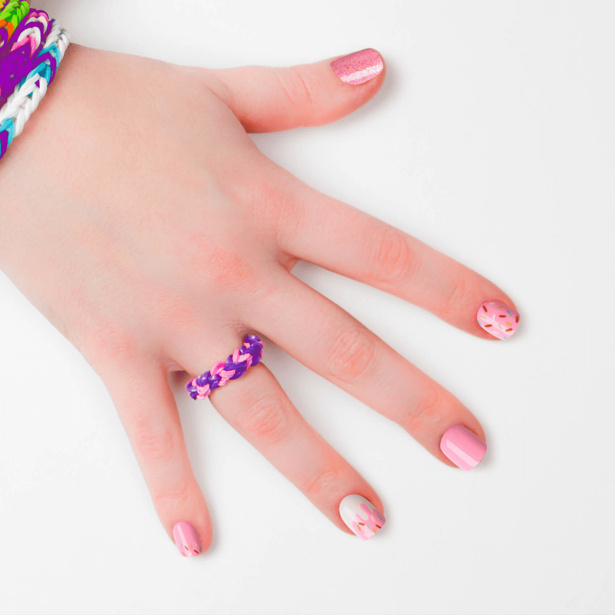 Hand with five press-on nails in pink tones, featuring sprinkles and dripping icing design. Hand wears colorful woven bracelets and ring on white background.