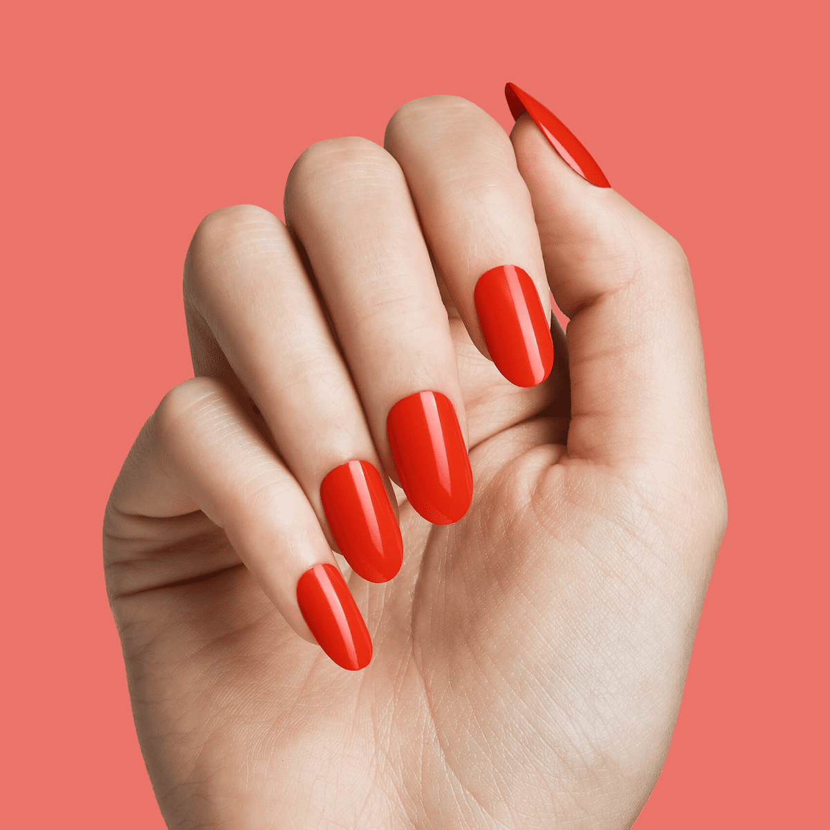 Hand with neatly manicured, oval-shaped full-cover nails painted in vibrant red, shown against a coral background.