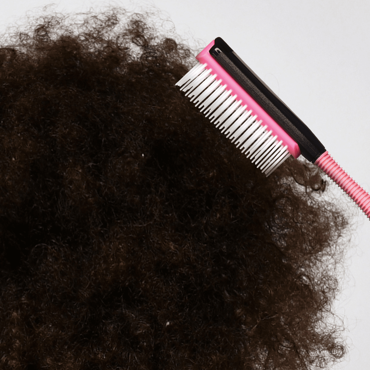 Pink mini detangling brush with white soft bristles and black non-slip grip, shown brushing thick, curly dark hair against a white background.
