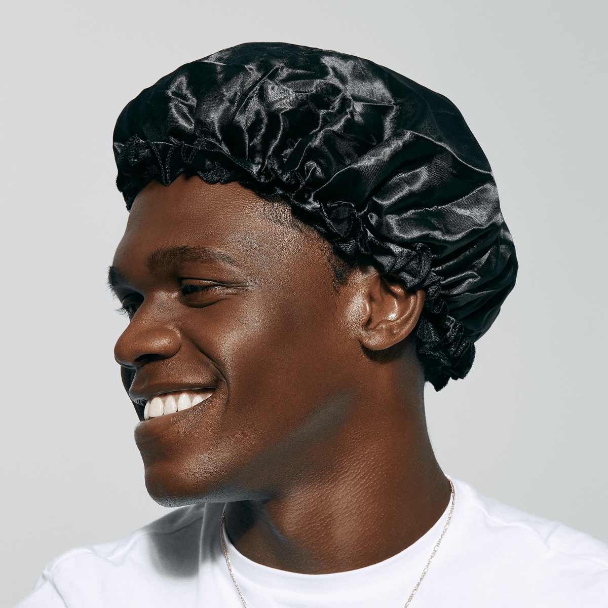 Smiling man wearing a black satin bonnet with gathered elastic edge, shown in close-up profile against a plain light gray background.