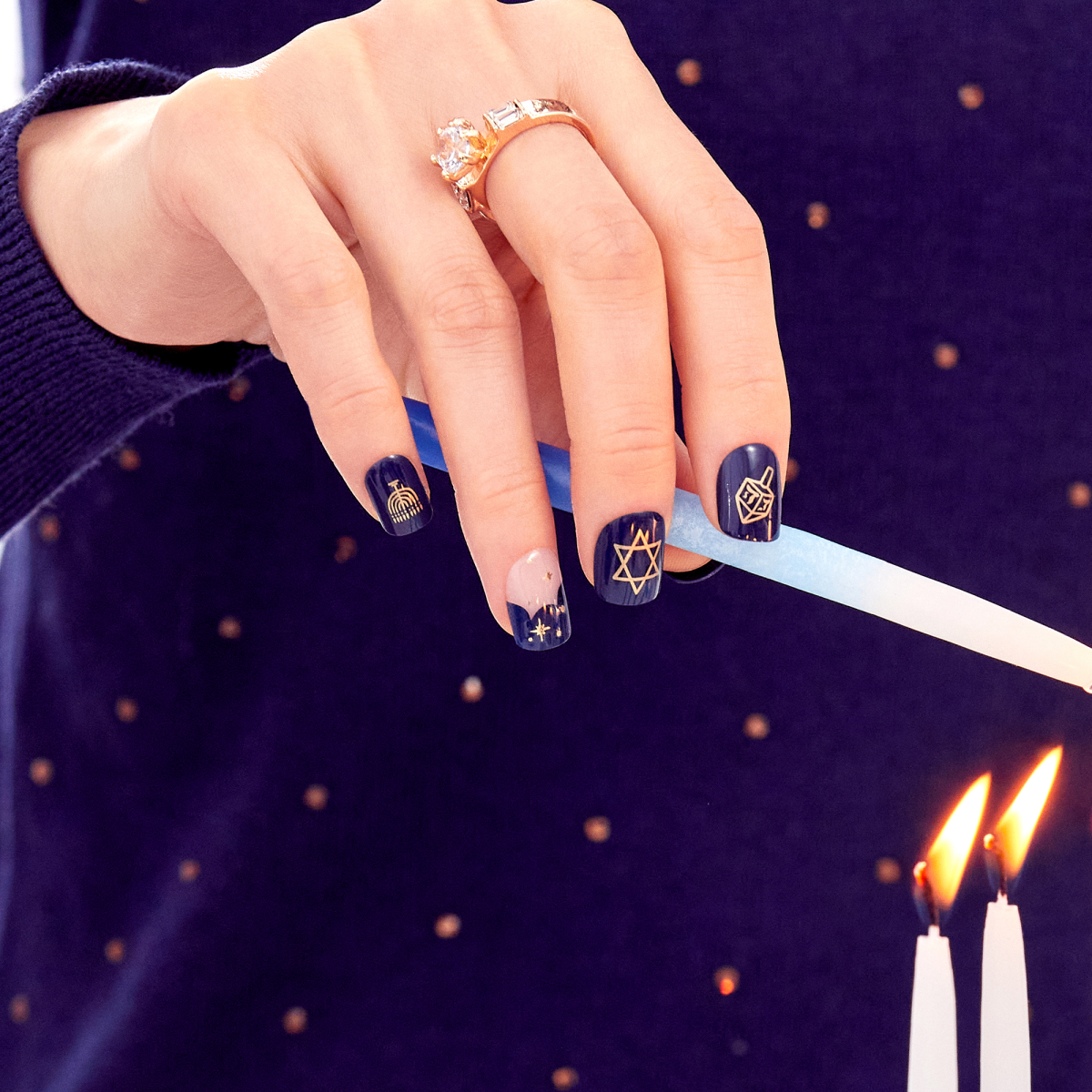 Hand with navy blue Hanukkah-themed press on nails showing gold designs of a menorah, dreidel, Star of David, and gold sparkles, lighting a candle.