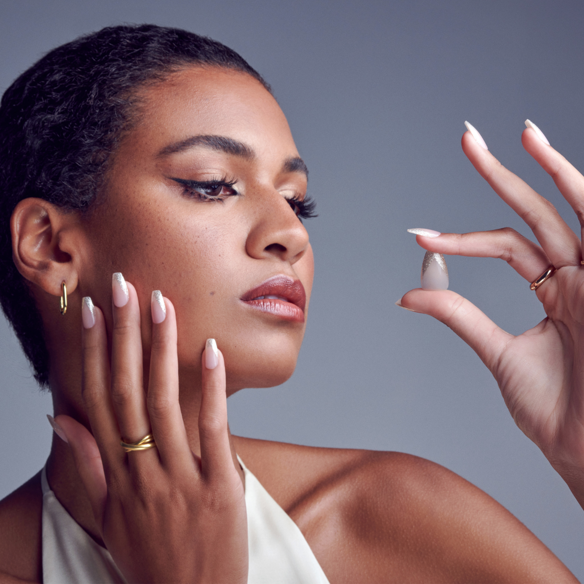 Woman with smooth skin and natural makeup holds a single almond-shaped, glitter-tipped soft gel nail, matching the nails on her hand.