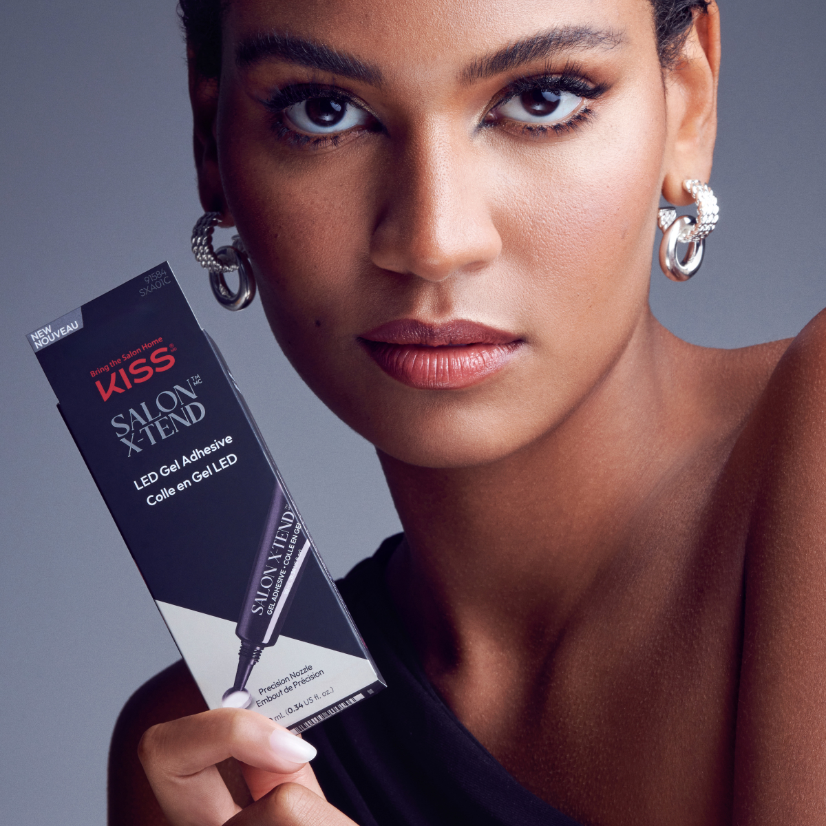 Woman with smooth skin and silver hoop earrings holding KISS Salon X-tend LED Gel Adhesive packaging, showing the precision nozzle and bilingual product name.