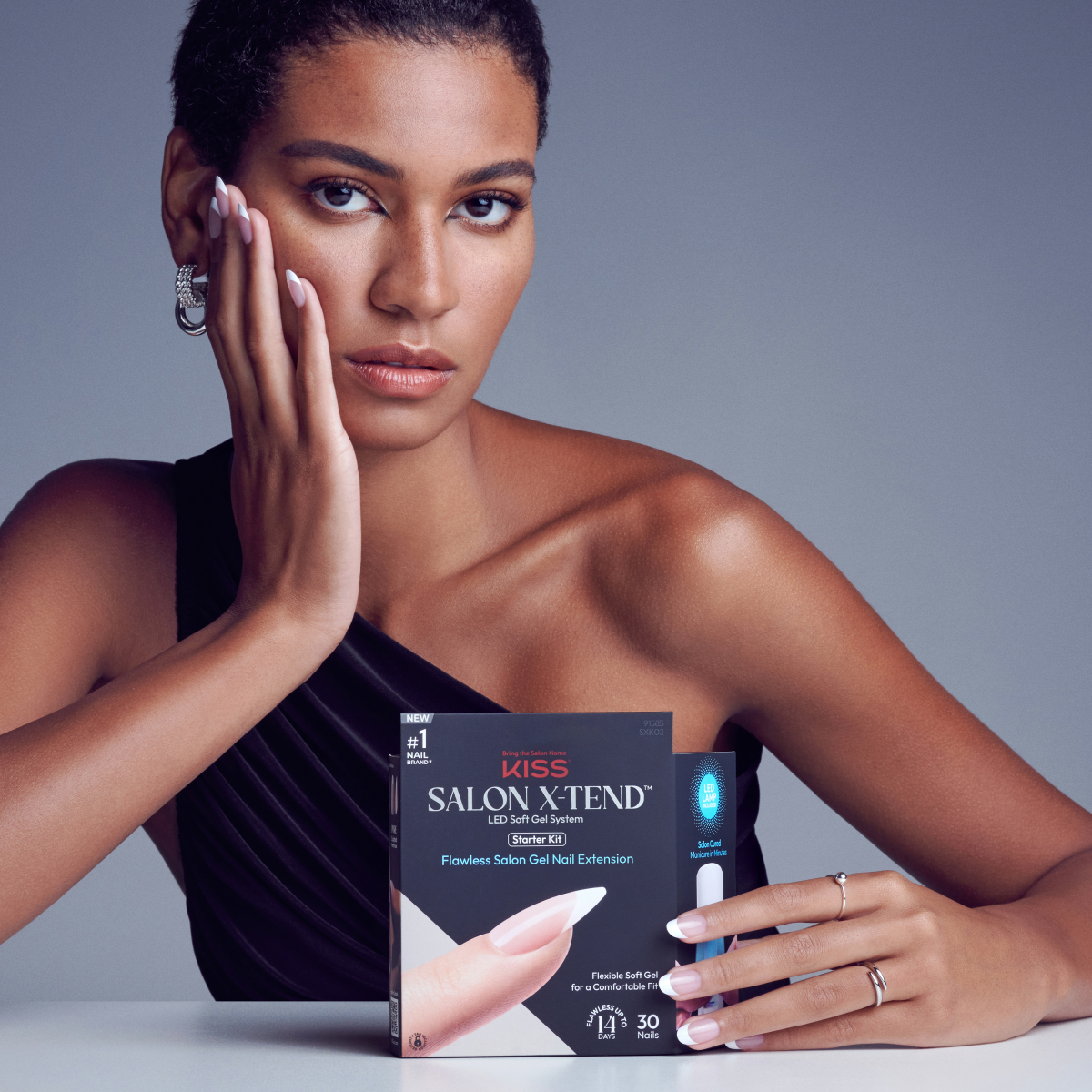 Woman with medium skin tone and short hair wears a black one-shoulder top, showing off long, soft gel French tip nails while holding the black KISS Salon X-tend LED Soft Gel Starter Kit box.