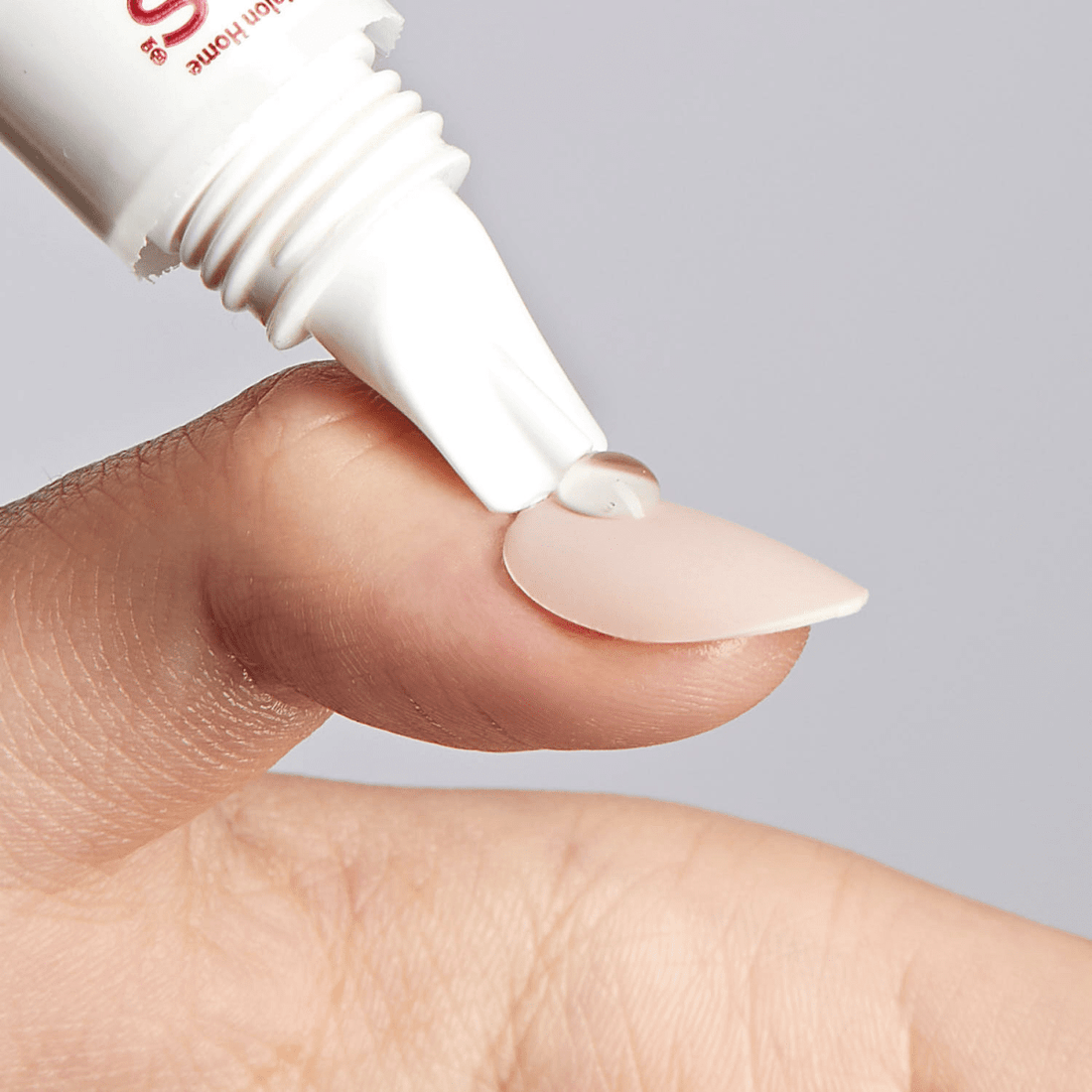 Clear gel being applied from a white tube onto a light pink press-on nail held on a fingertip, demonstrating nail glue remover use.