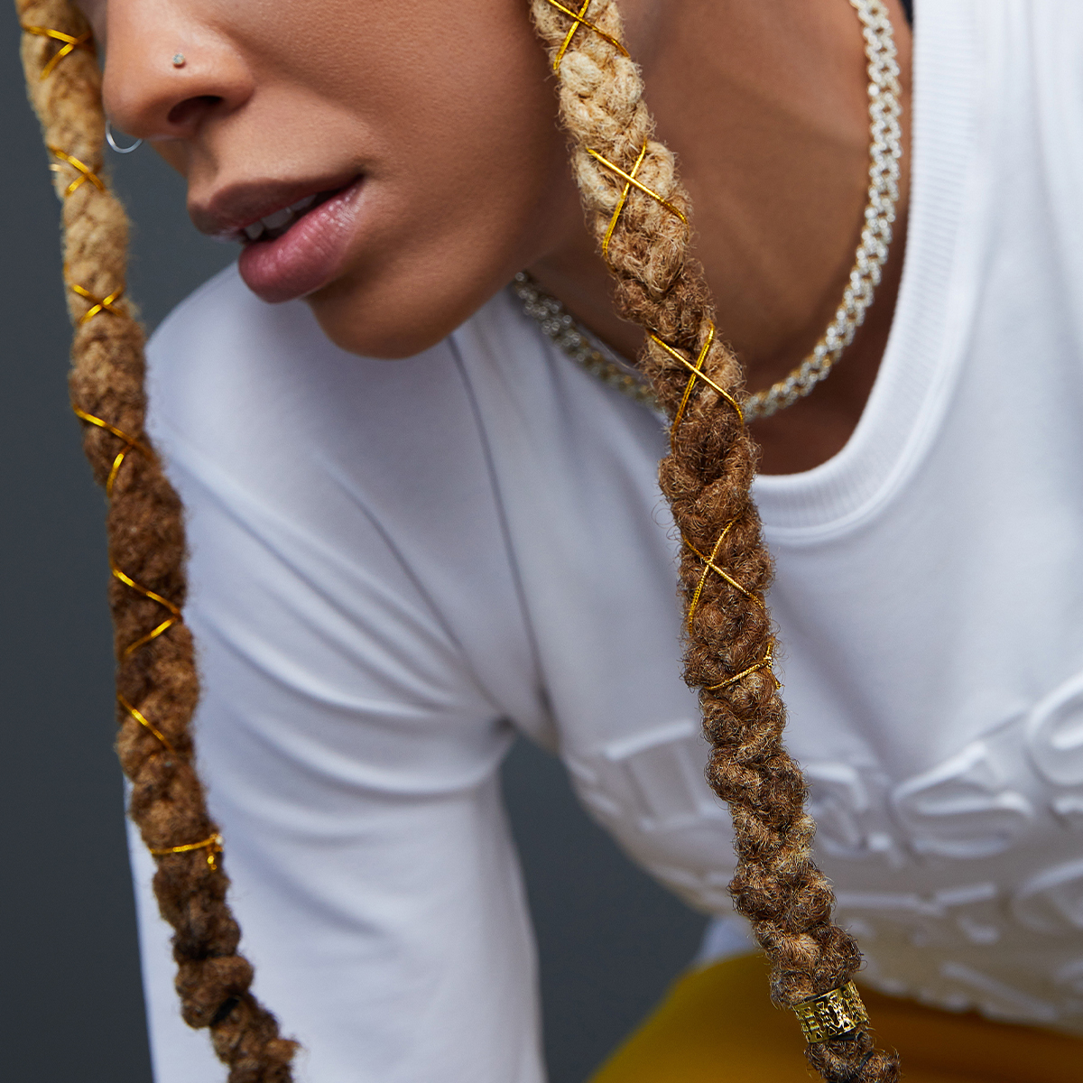 Close-up of two blonde to brown braided dreadlocks decorated with shiny gold spiral braid charms on a person wearing a white shirt and silver chain necklace.