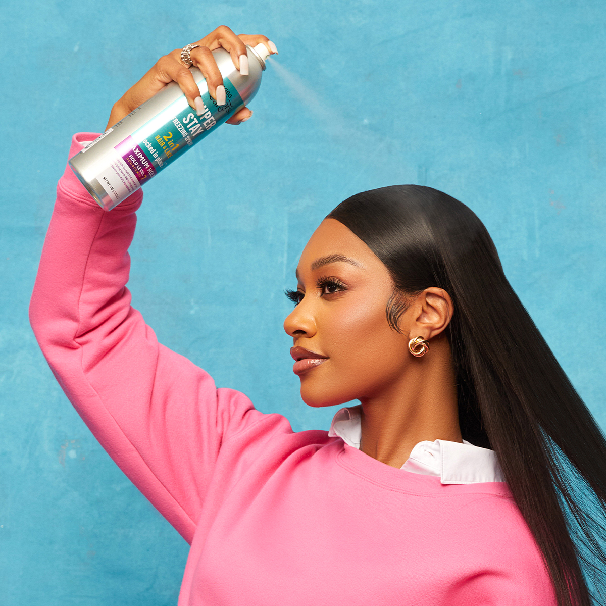 Woman wearing pink sweatshirt sprays KISS Super Stay Freezing Spray Maximum Hold from a silver can with blue and purple label against blue background.