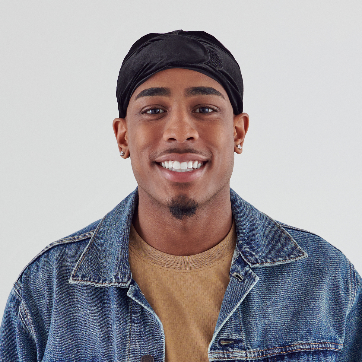 Young man wearing a black velvet durag tied at the back, smiling, dressed in a tan shirt and blue denim jacket against a plain light background.