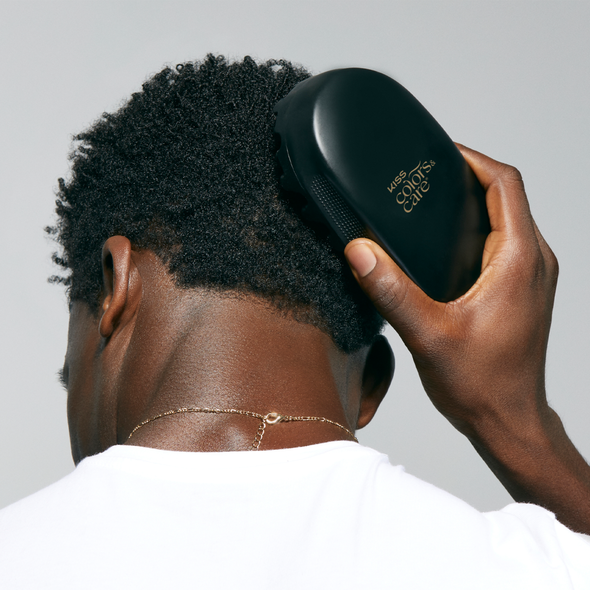 Man with short curly black hair wearing a white shirt, using a black KISS Colors &amp; Care hair brush on the back of his head.
