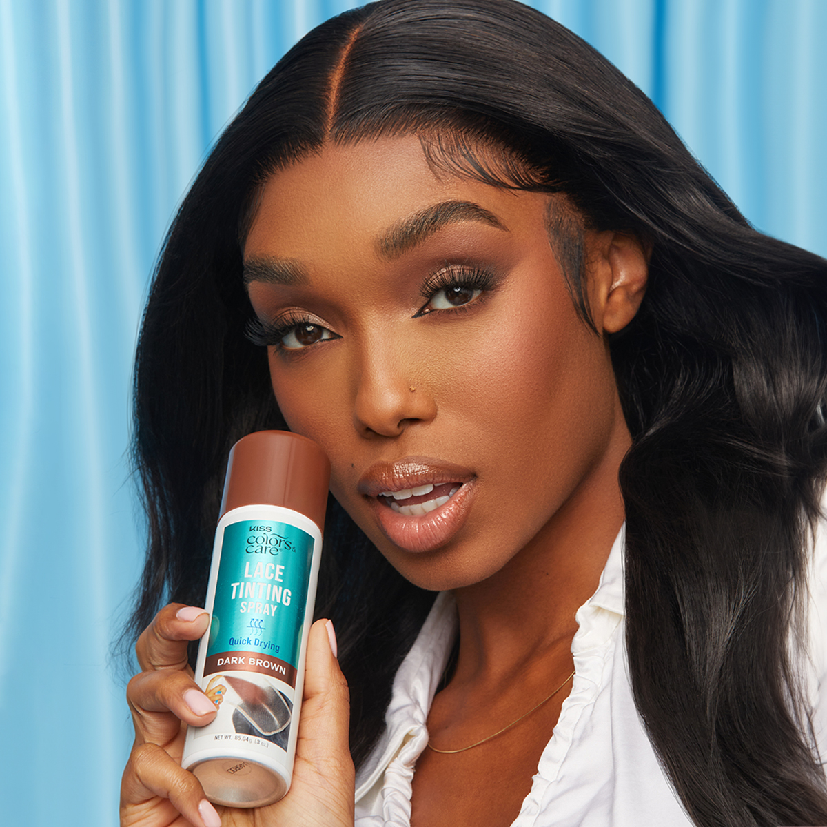 Woman with long dark brown hair holding KISS Colors &amp; Care Lace Tinting Spray in dark brown, labeled "Quick Drying" on a blue background.